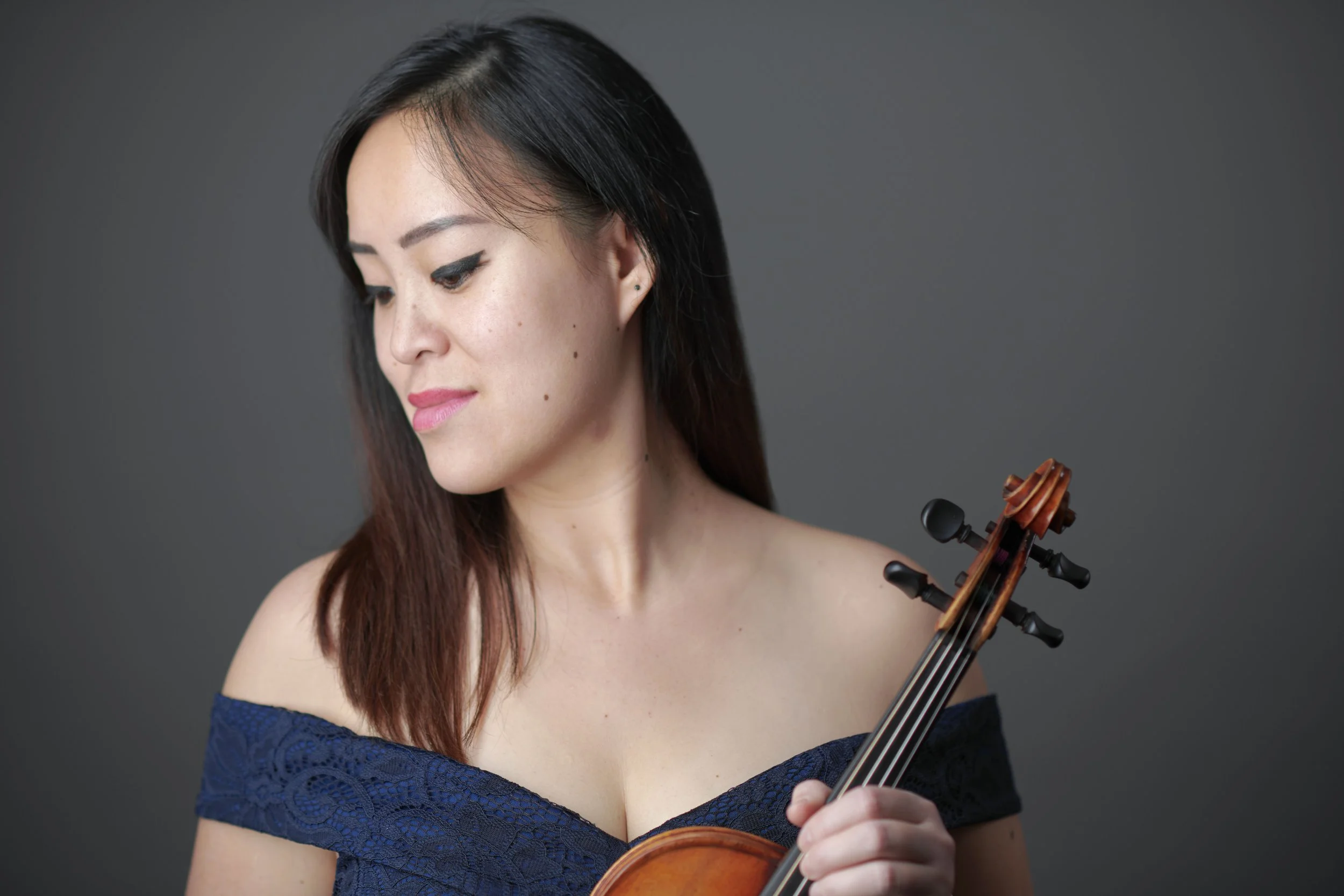 Woman in off-shoulder blue dress holding a violin against a gray background.