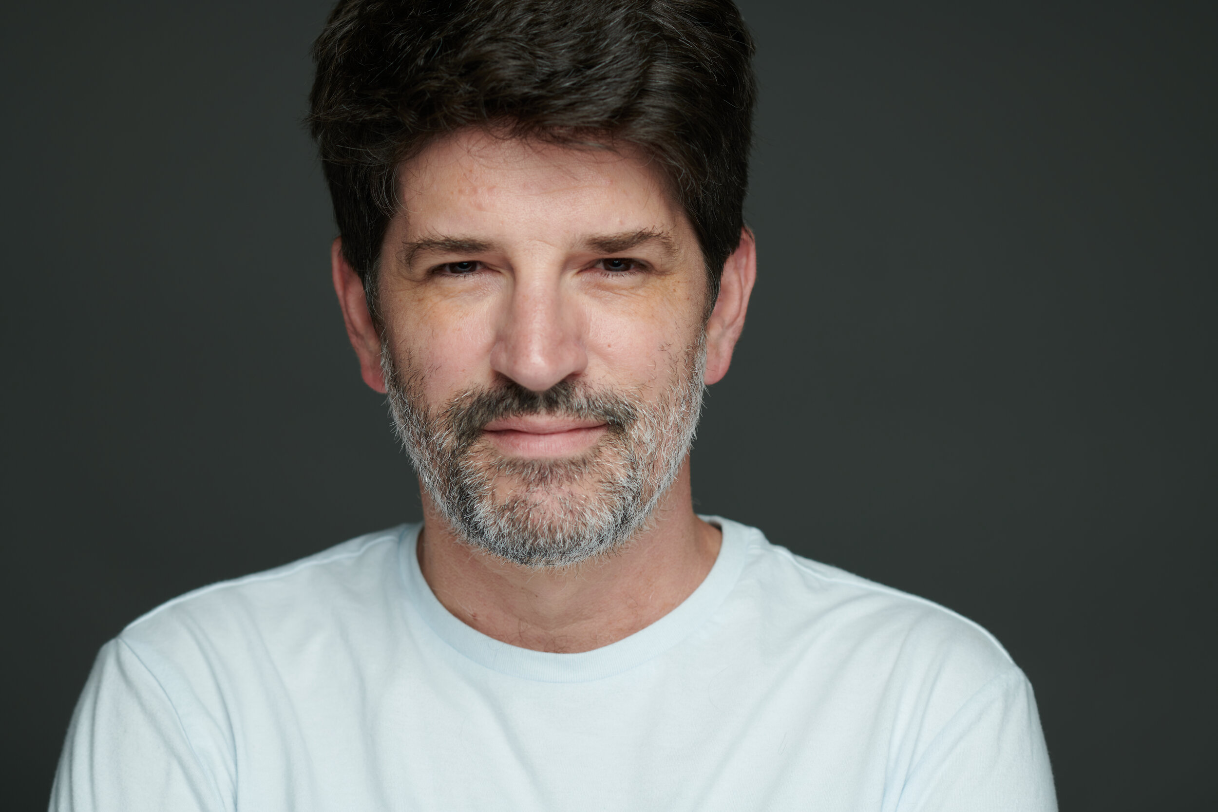 Portrait of a middle-aged man with dark hair and beard, wearing a white shirt, against a dark gray background.