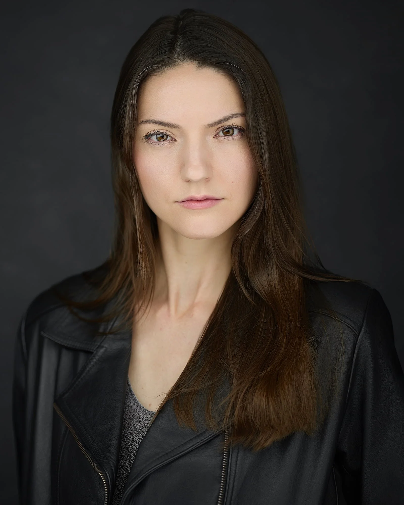 Theatrical headshot of an actress wearing a grey sweater and black leather jacket against a black backdrop
