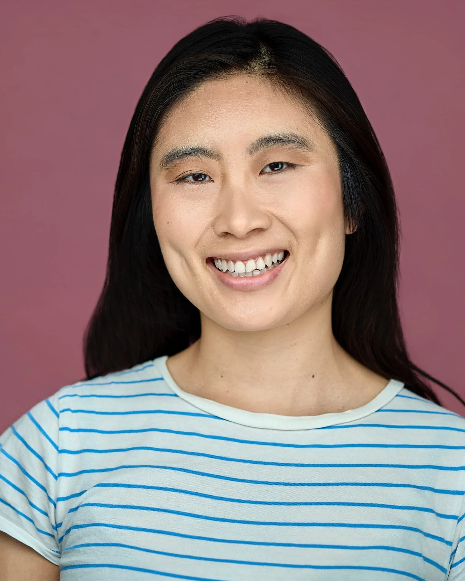 Actress headshot of an Asian woman wearing a blue and white striped shirt against a pink backdrop