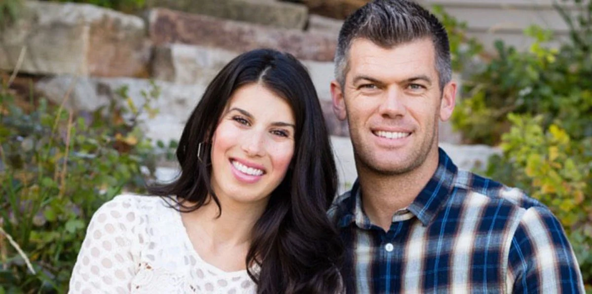 A smiling woman with long dark hair and a man with short dark hair stand outdoors with greenery and a brick wall in the background.