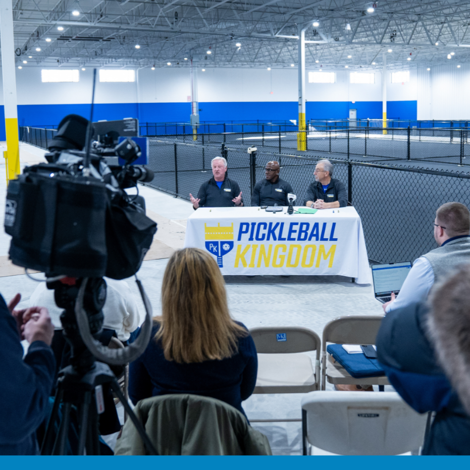Press conference at Pickleball Kingdom indoors, with three speakers at a table covered with a Pickleball Kingdom banner, surrounded by media personnel and cameras, empty pickleball courts in the background.