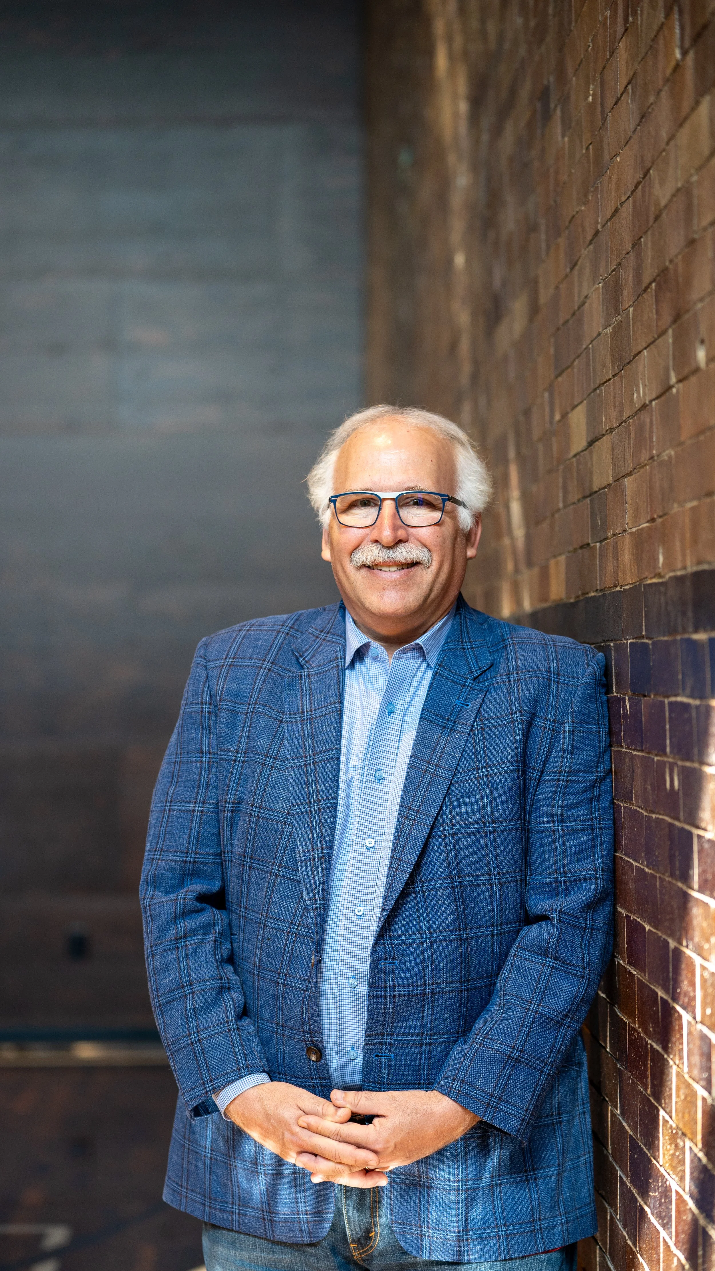 A smiling elderly man with white hair and glasses, wearing a blue checked blazer and light blue shirt, standing with hands clasped in front of them against a brick wall in an indoor setting.