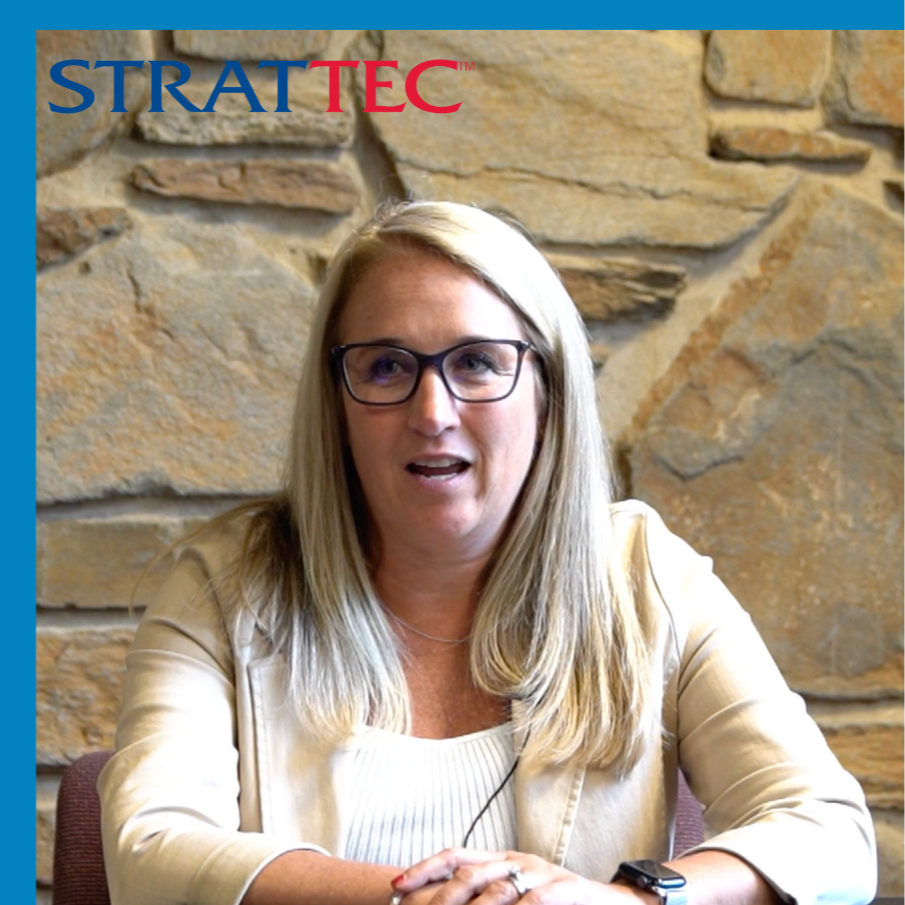 Side-by-side of two people in a meeting room with stone walls. The woman on the left has long blonde hair, glasses, a beige blazer, and is speaking. The man on the right has short hair, glasses, a blue checkered shirt, and is gesturing with his hand. Subtitles are present at the bottom of both sides of the image.