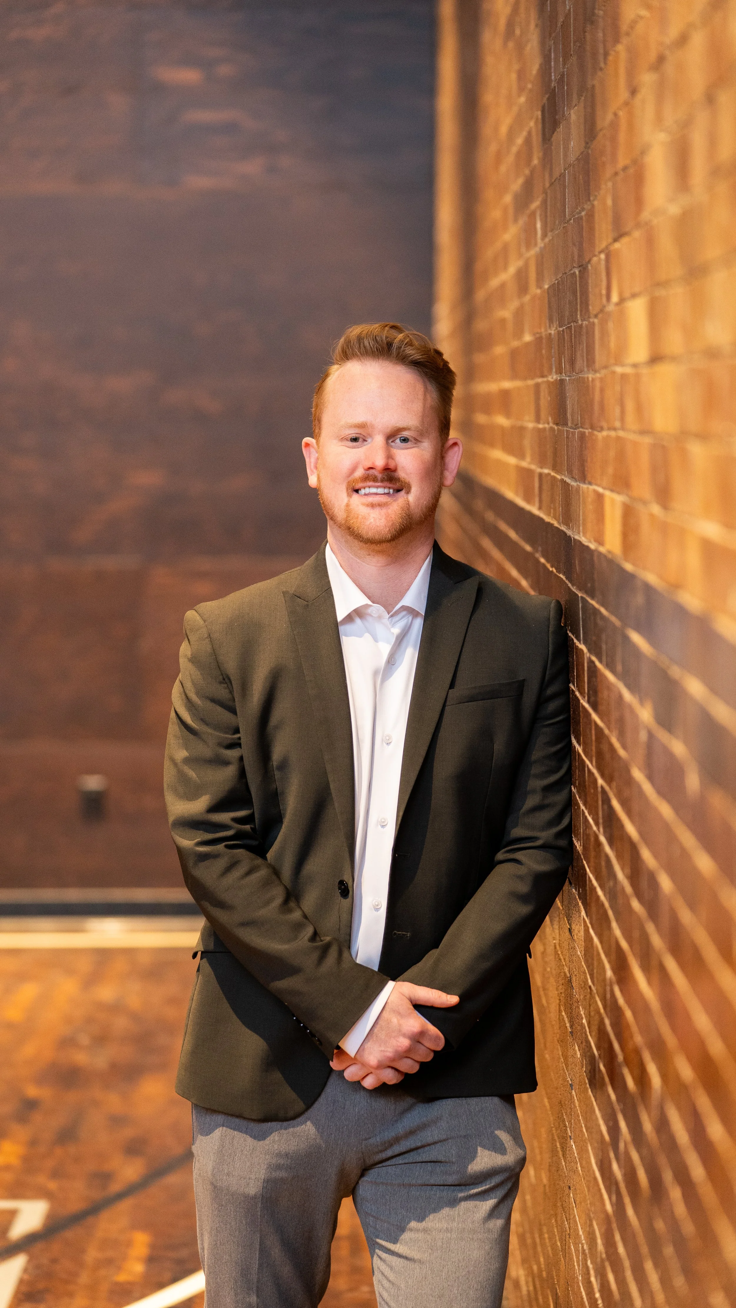 A man in a black suit jacket, white shirt, and gray pants leaning against a brick wall in an indoor setting.