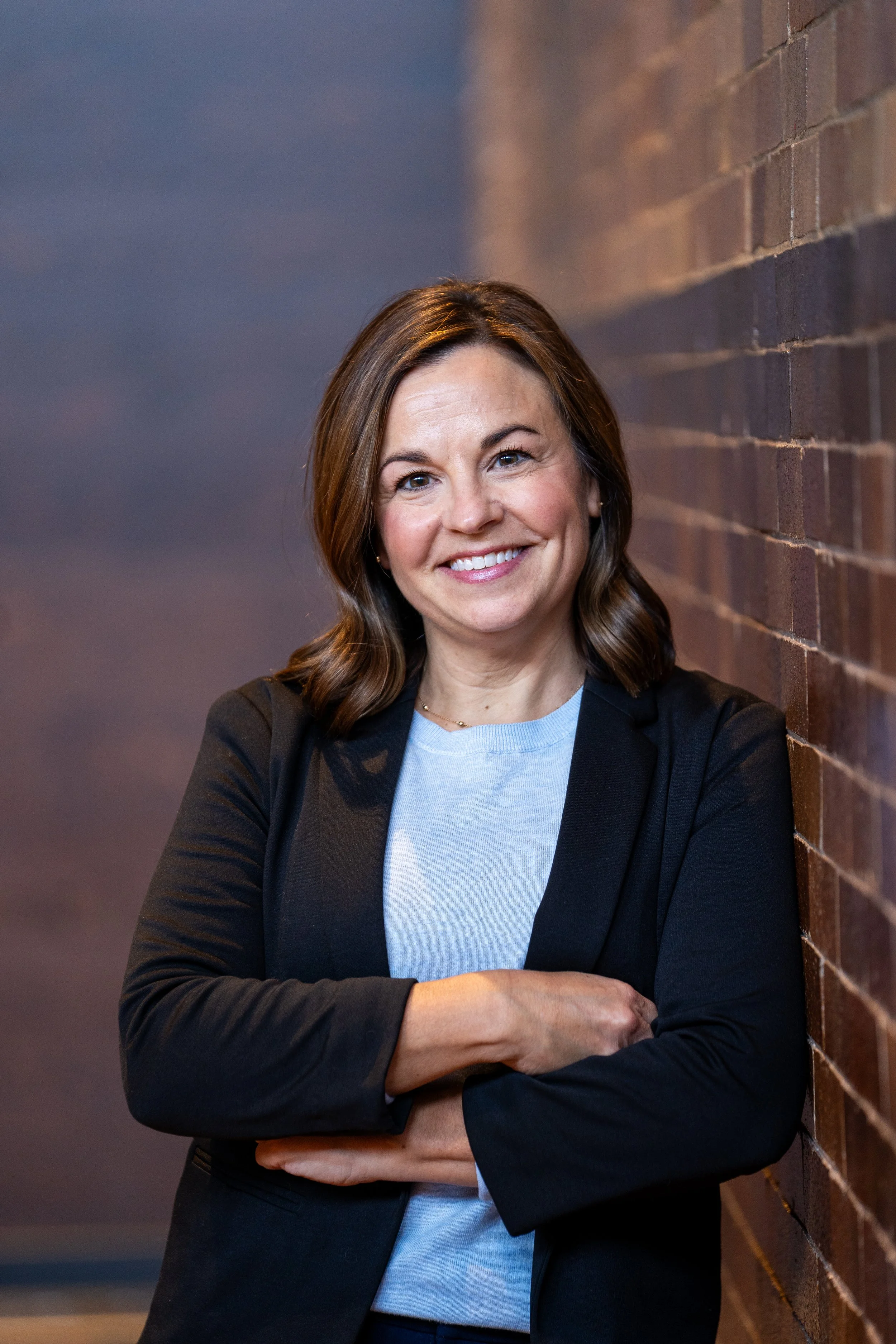 A smiling woman with shoulder-length brown hair, wearing a black blazer over a light blue shirt, leaning against a brick wall.