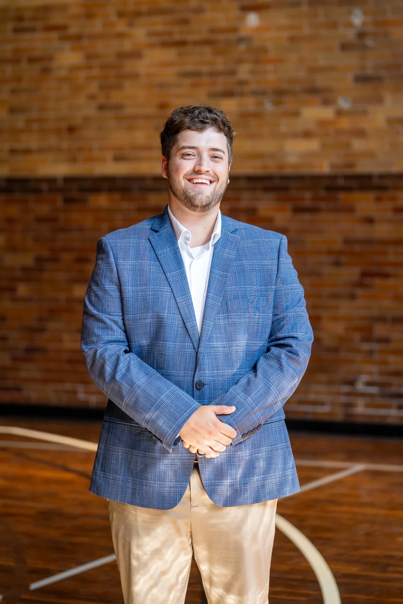 A man with a beard and short brown hair wearing a blue checkered blazer and light-colored pants standing in front of a brick wall, smiling and looking at the camera.