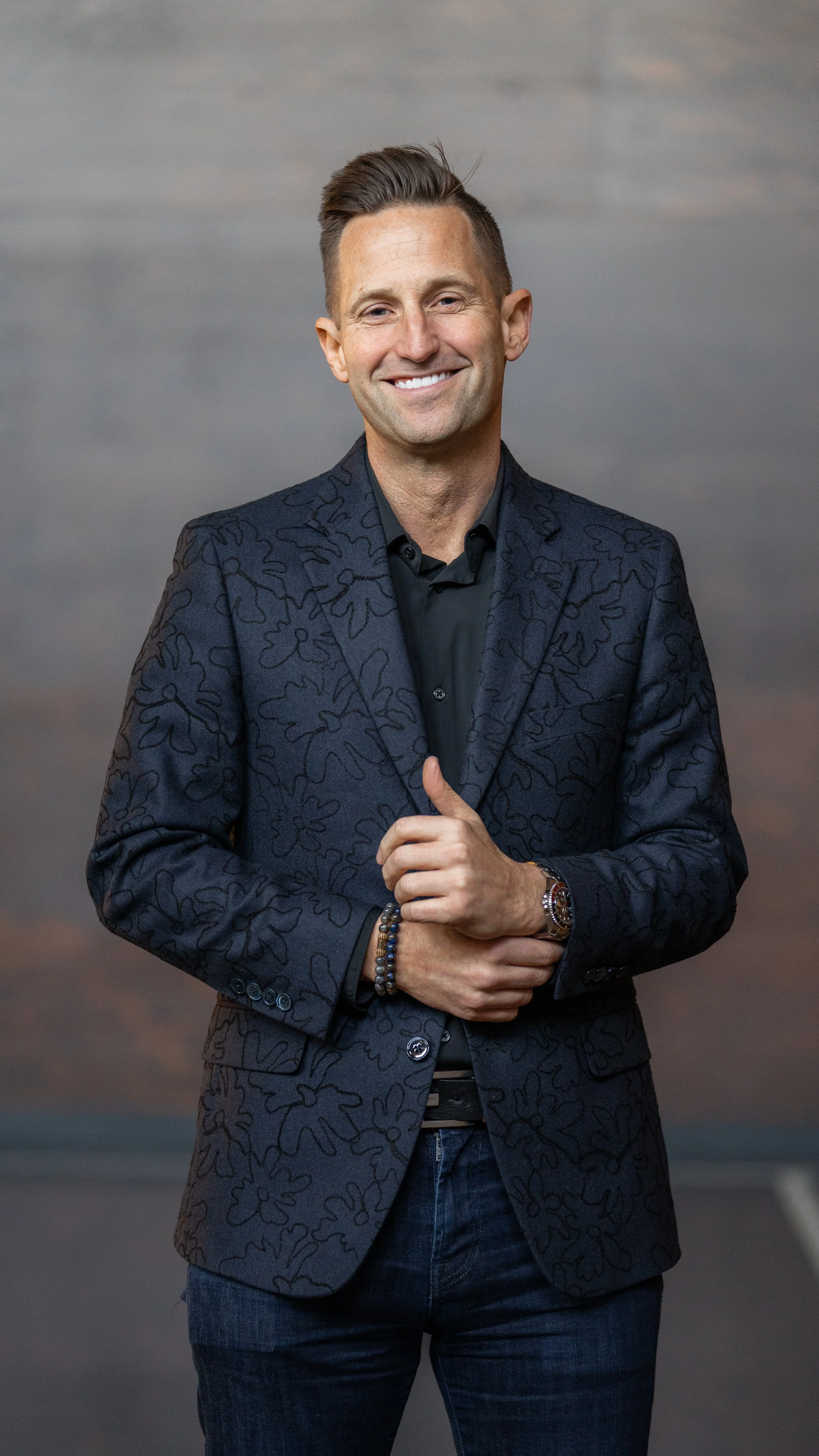 A smiling man in a patterned dark blazer and black shirt standing outdoors during sunset.