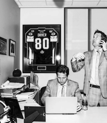 Two men in an office, one sitting at a desk working on a laptop, the other standing using a phone. Framed sports jersey of a Giants player with the number 80 is on the wall behind them.