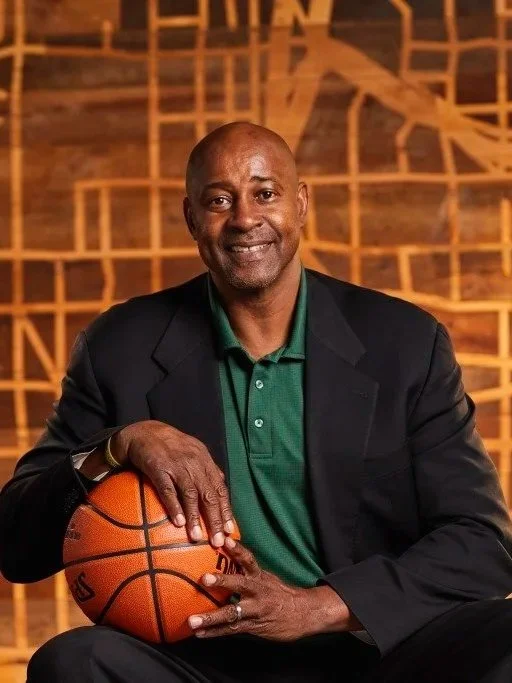 A man in a black suit and green polo shirt sitting with a basketball in his lap, in front of a wooden geometric background.