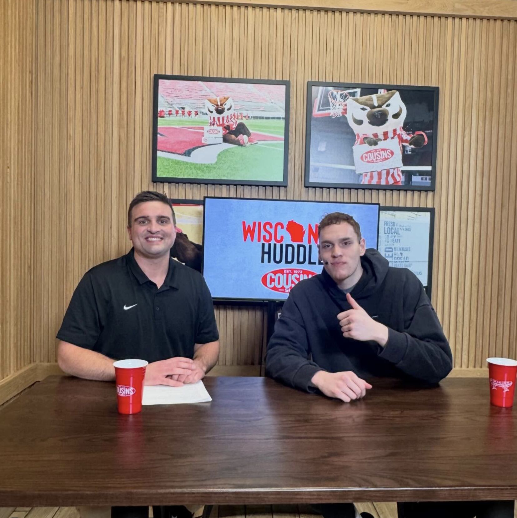 Two men sitting at a wooden table with red cups in front of them, in a room with wooden panel walls and TV screens showing Wisconsin mascot images and a logo for Wisconsin Huddles.
