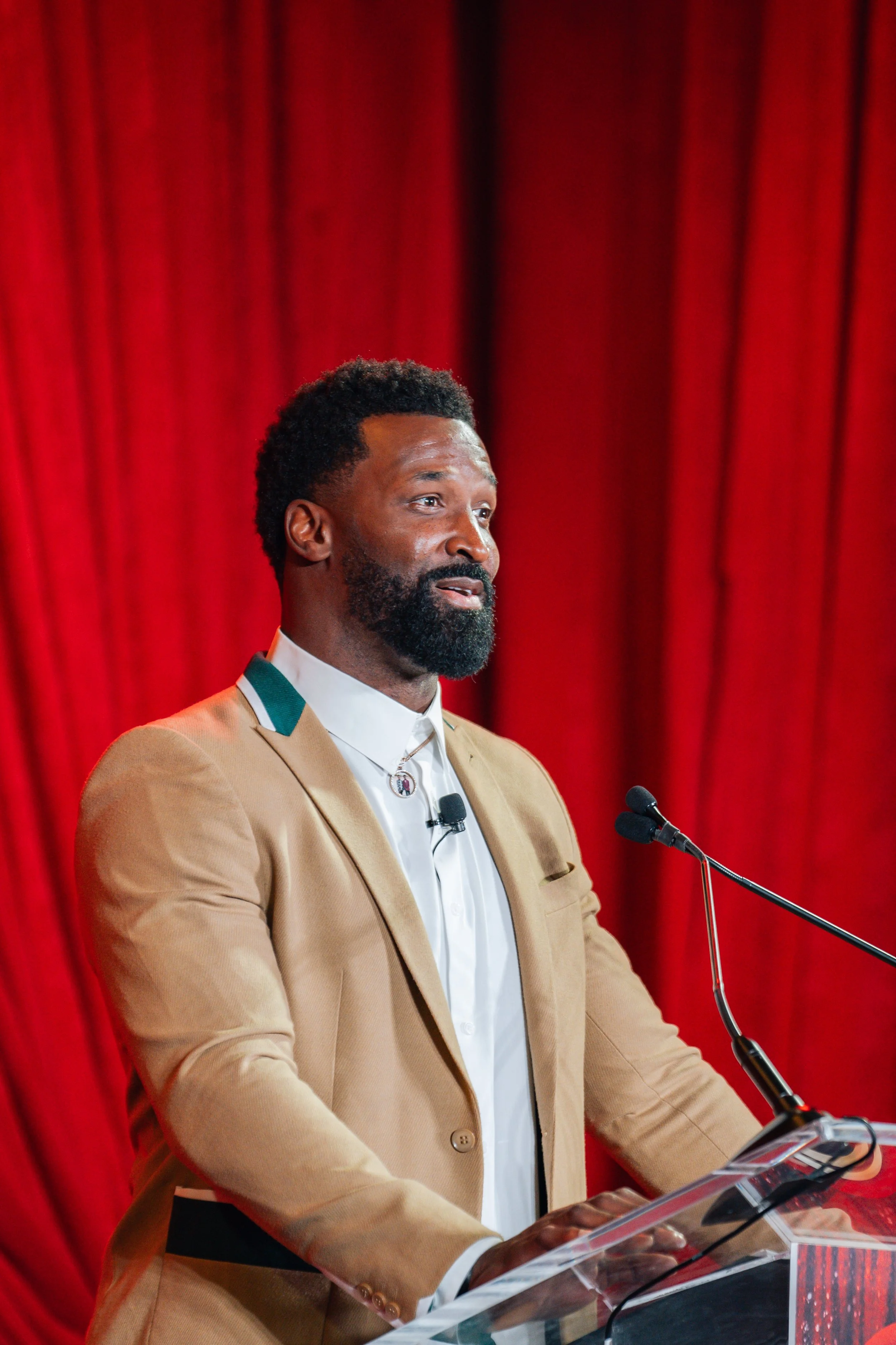 A man with dark hair and a beard standing at a podium, speaking into a microphone, with a red curtain in the background.