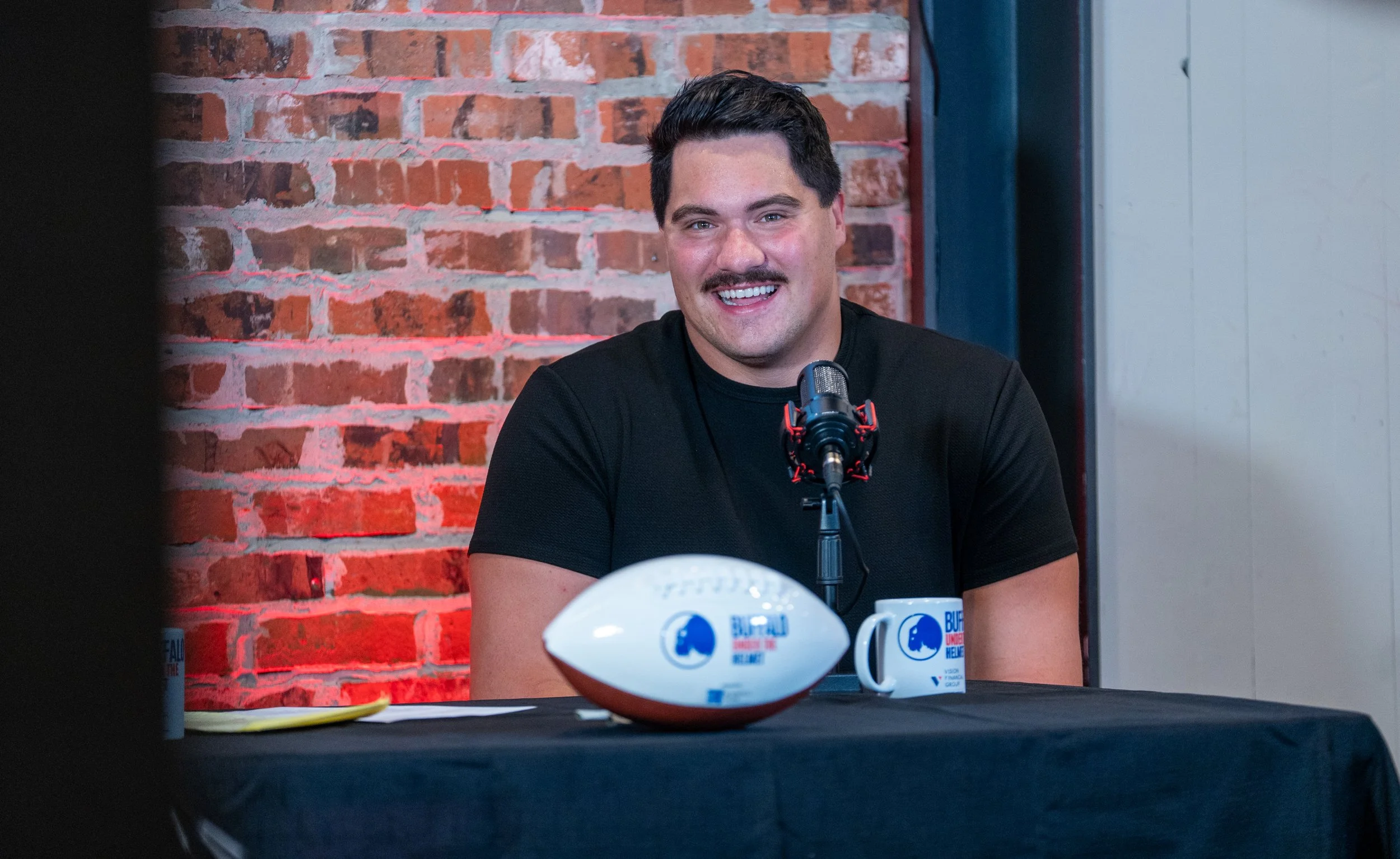 A man with dark hair and a mustache, smiling, sits at a table with a microphone, a football, and a mug that has the Buffalo logo and the words 'Buffalo Under the Helmet' during a podcast or interview. The background is a red brick wall.
