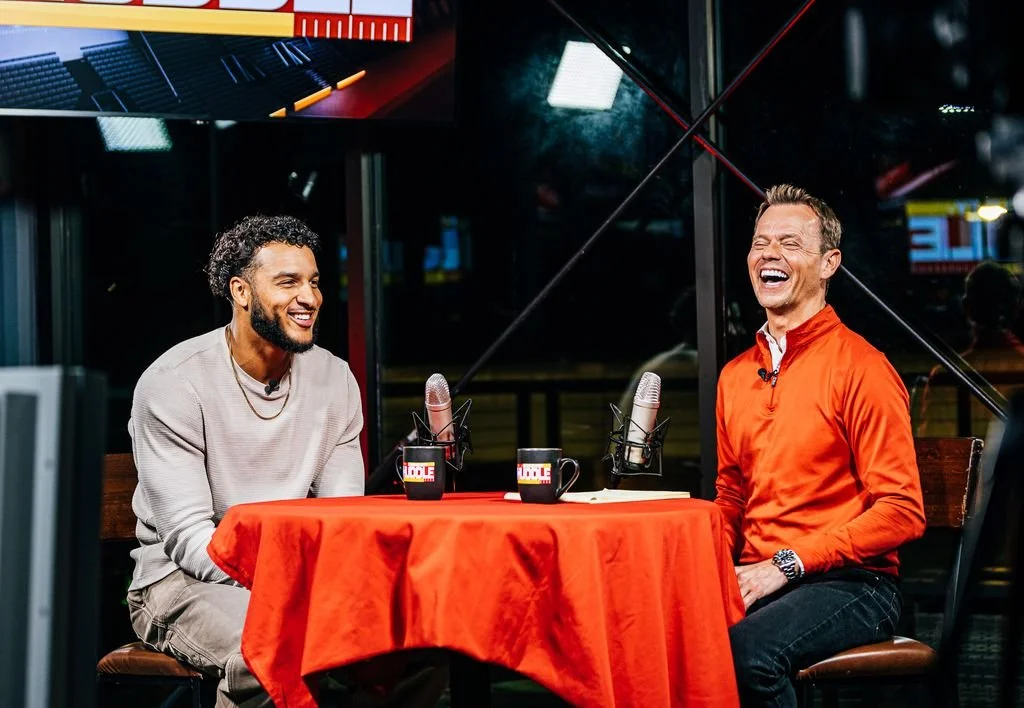 Two men sitting at a table with orange tablecloth, microphones, and coffee mugs, laughing and having a conversation in a studio.