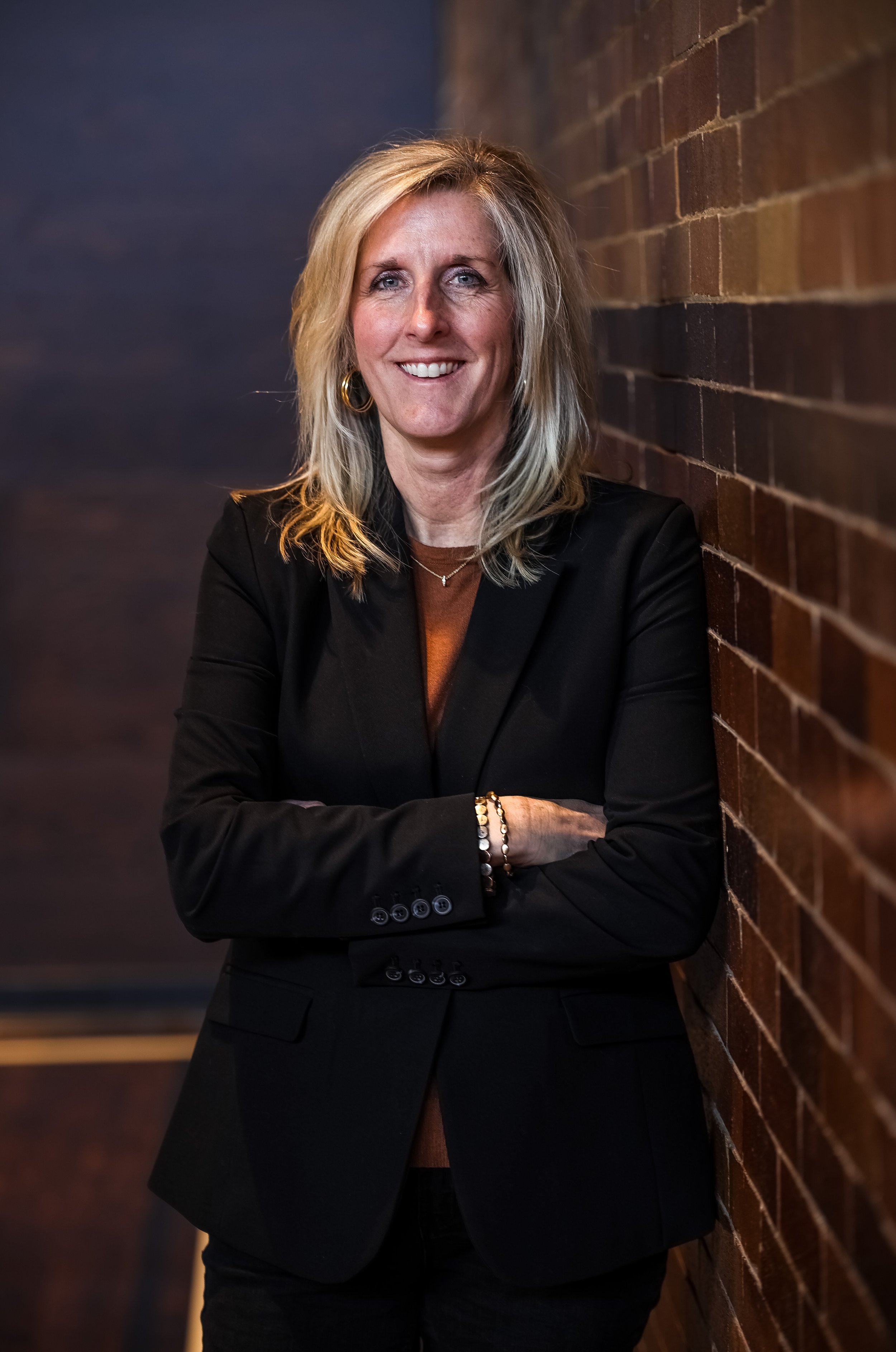A woman with shoulder-length blonde hair, wearing a black blazer, standing with arms crossed and smiling, against a brick wall background.