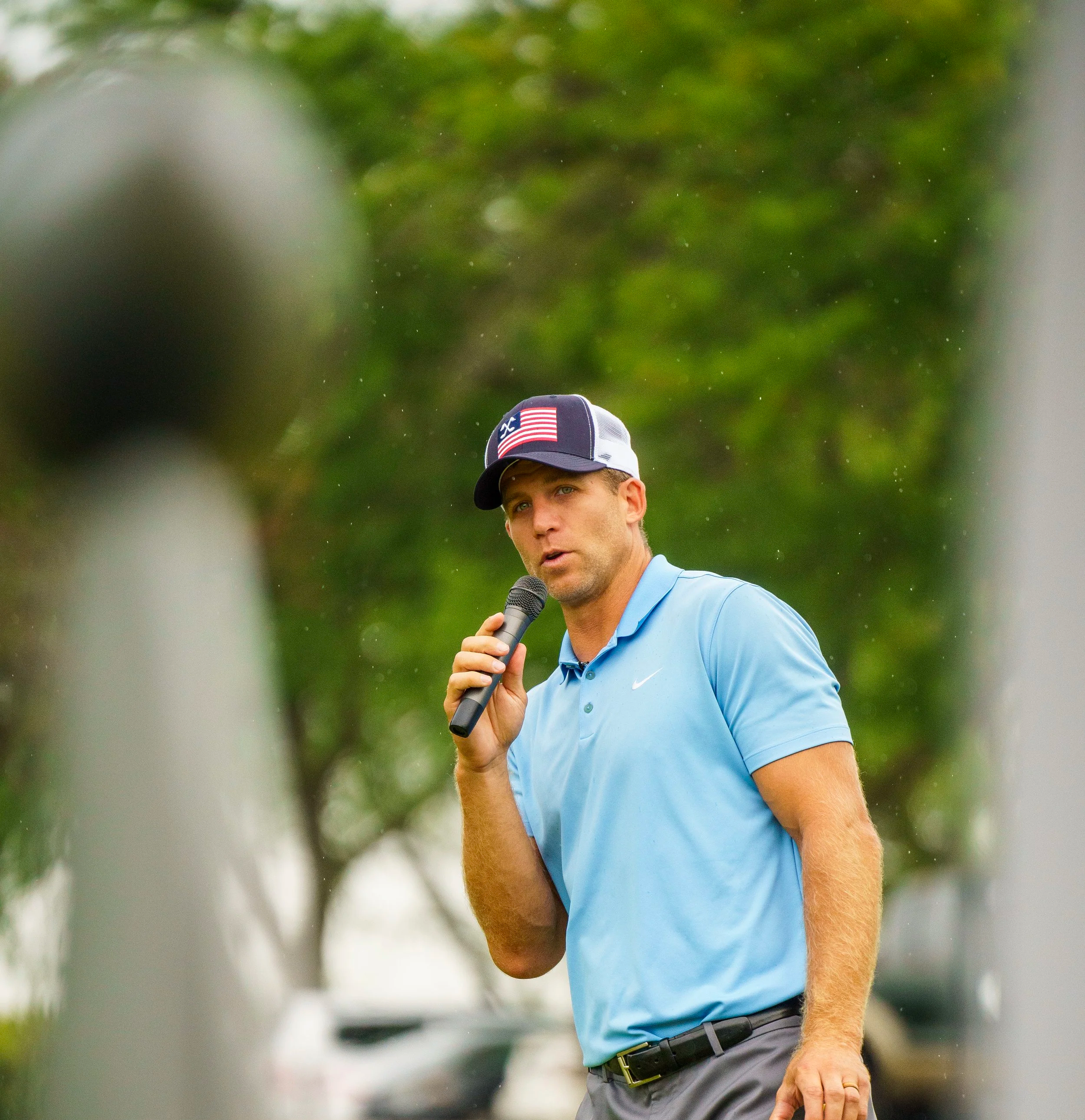 A man in a light blue polo shirt and a baseball cap holding a microphone, speaking outdoors with trees and blurred vehicles in the background, seen through a narrow frame.