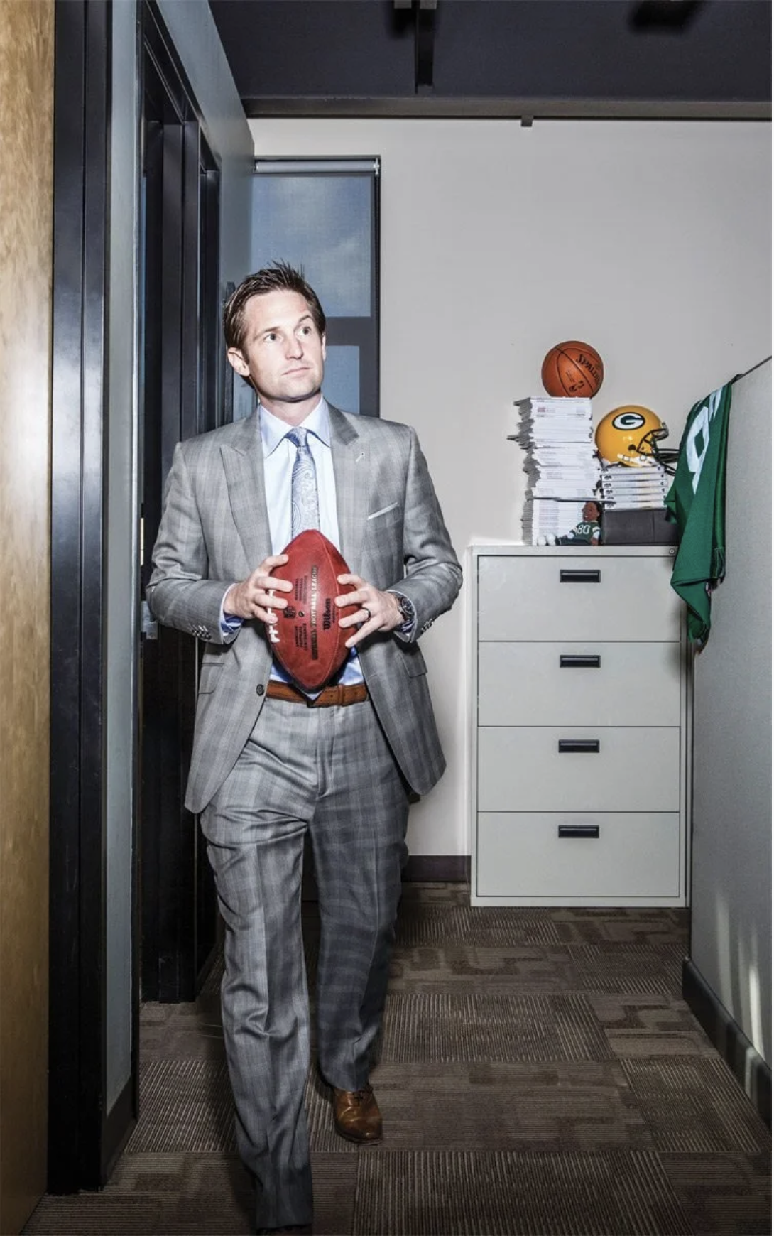A man in a gray plaid suit holding a football, standing in an office hallway with a stack of papers on a white filing cabinet, a basketball, a football helmet, and a Green Bay Packers jersey on top of the cabinet.