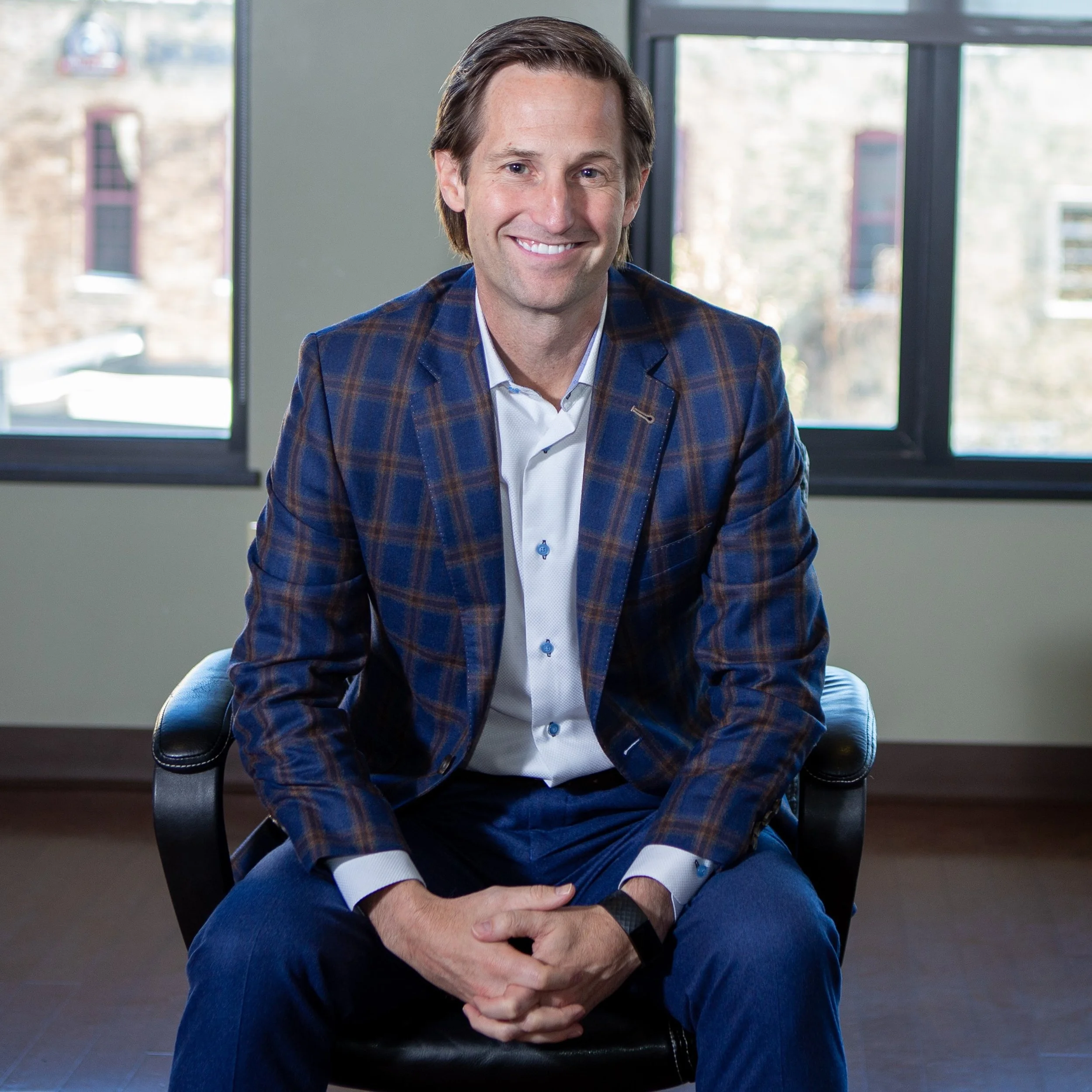 A man with light skin, brown hair, wearing a blue plaid blazer and white shirt, sitting on a black chair in front of a window with natural light.