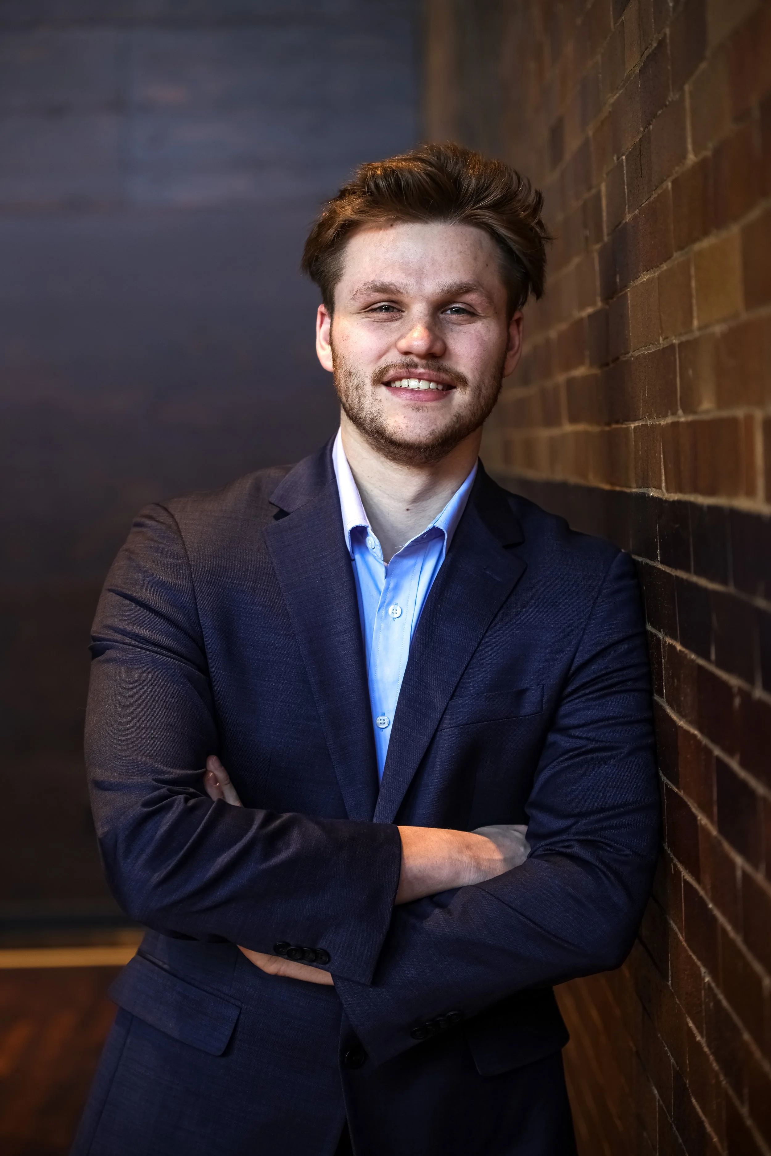 A young man with light brown hair, beard, and light skin, dressed in a blue suit and light blue shirt, standing with arms crossed, smiling, against a brick wall background.