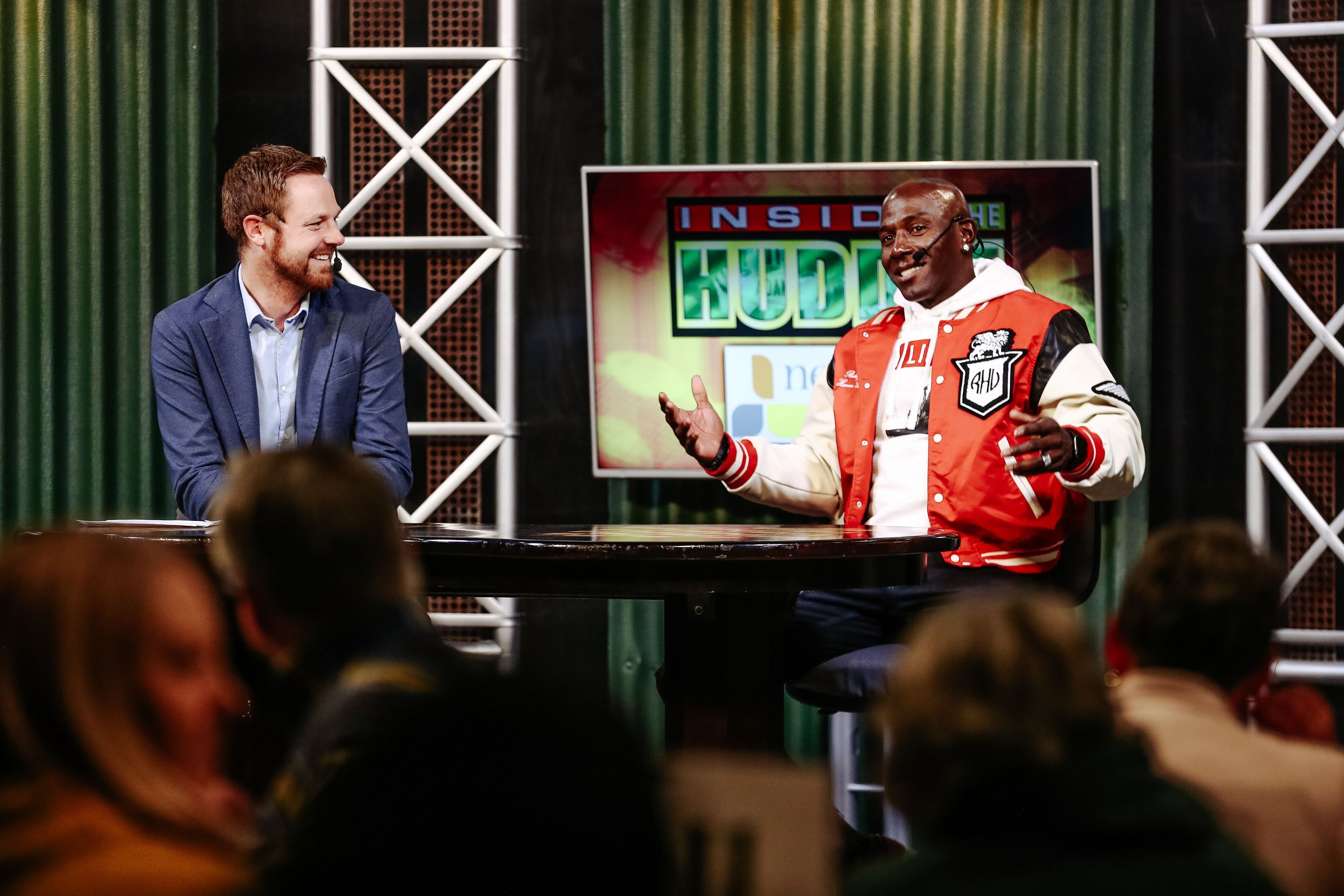 Two men are sitting at a round table on stage, engaged in a conversation. The man on the left has short brown hair and a beard, wearing a blue blazer, while the man on the right is bald, wearing glasses, and dressed in a red and white varsity jacket. Behind them is a large screen displaying the words "Inside the HUD" and a green background, with stage lighting and green curtains on either side.