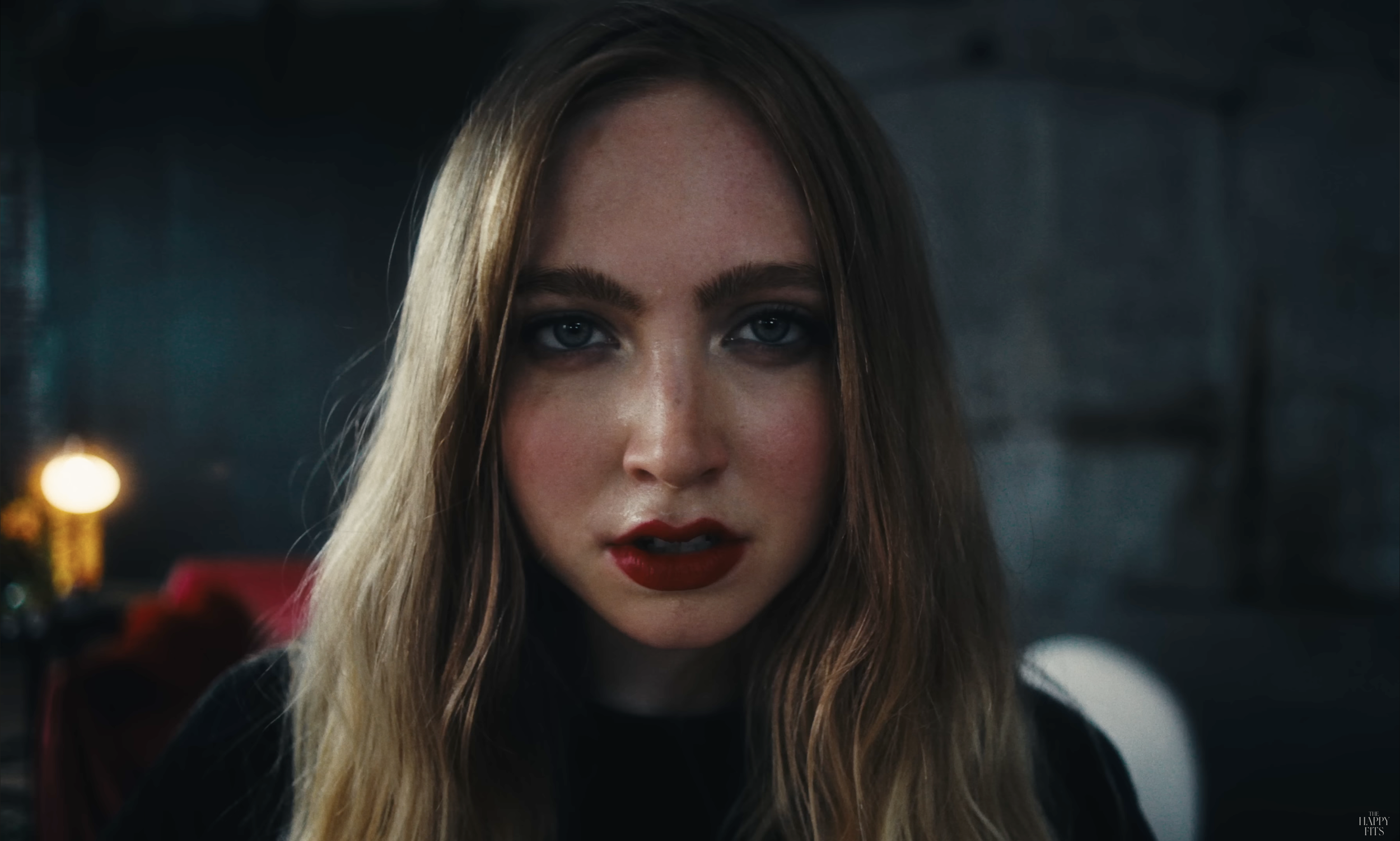 Close-up of a woman, Lucy Vally, with long blonde hair, blue eyes, wearing red lipstick, in a dimly lit room with blurred background.