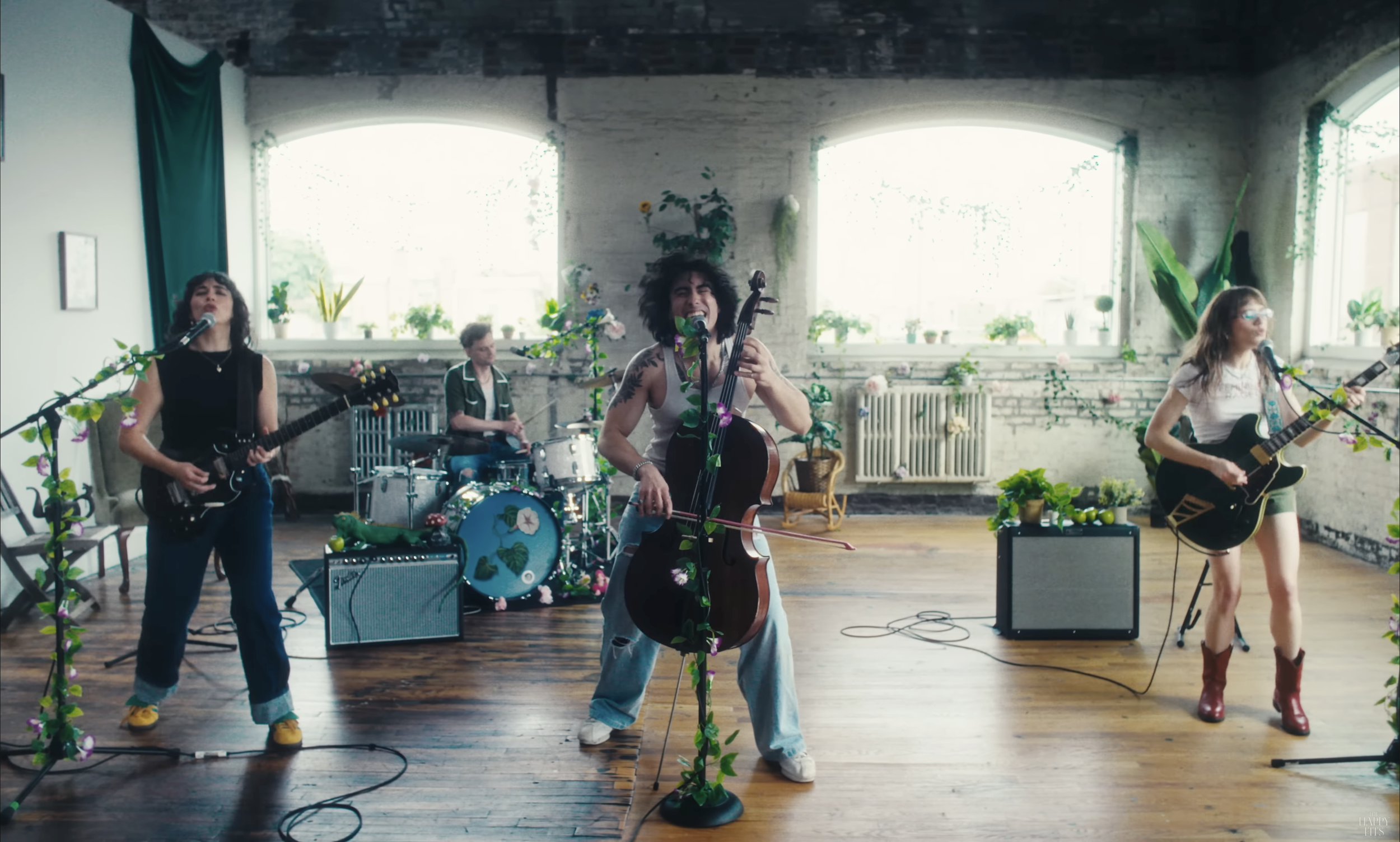 The Happy Fits performing with musical instruments in a bright room decorated with green plants and flowers, with large windows in the background.