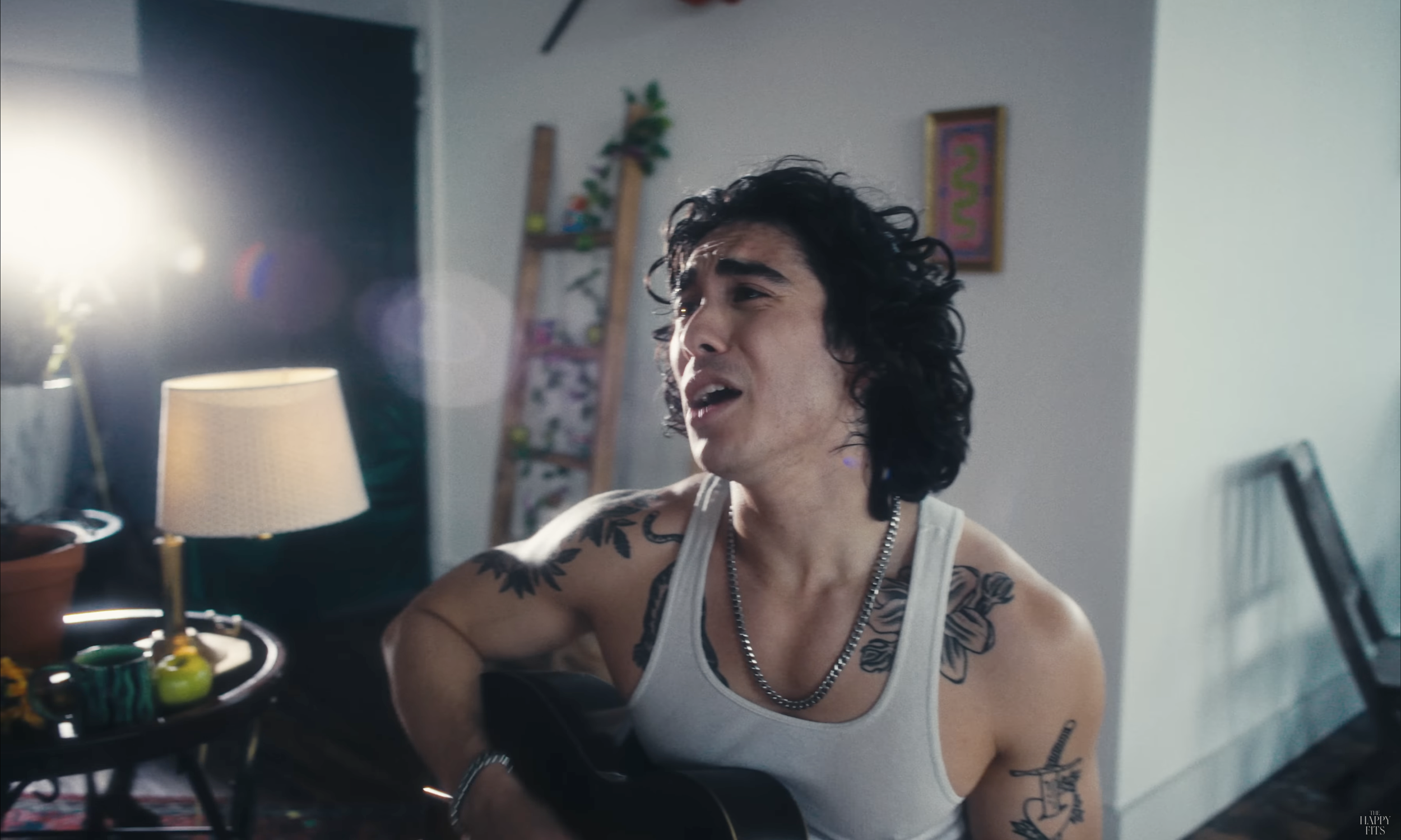 Calvin from The Happy Fits, a man with long, curly black hair and tattoos on his arms, wearing a white tank top and a silver chain, appears to be singing or playing guitar in a brightly lit room.