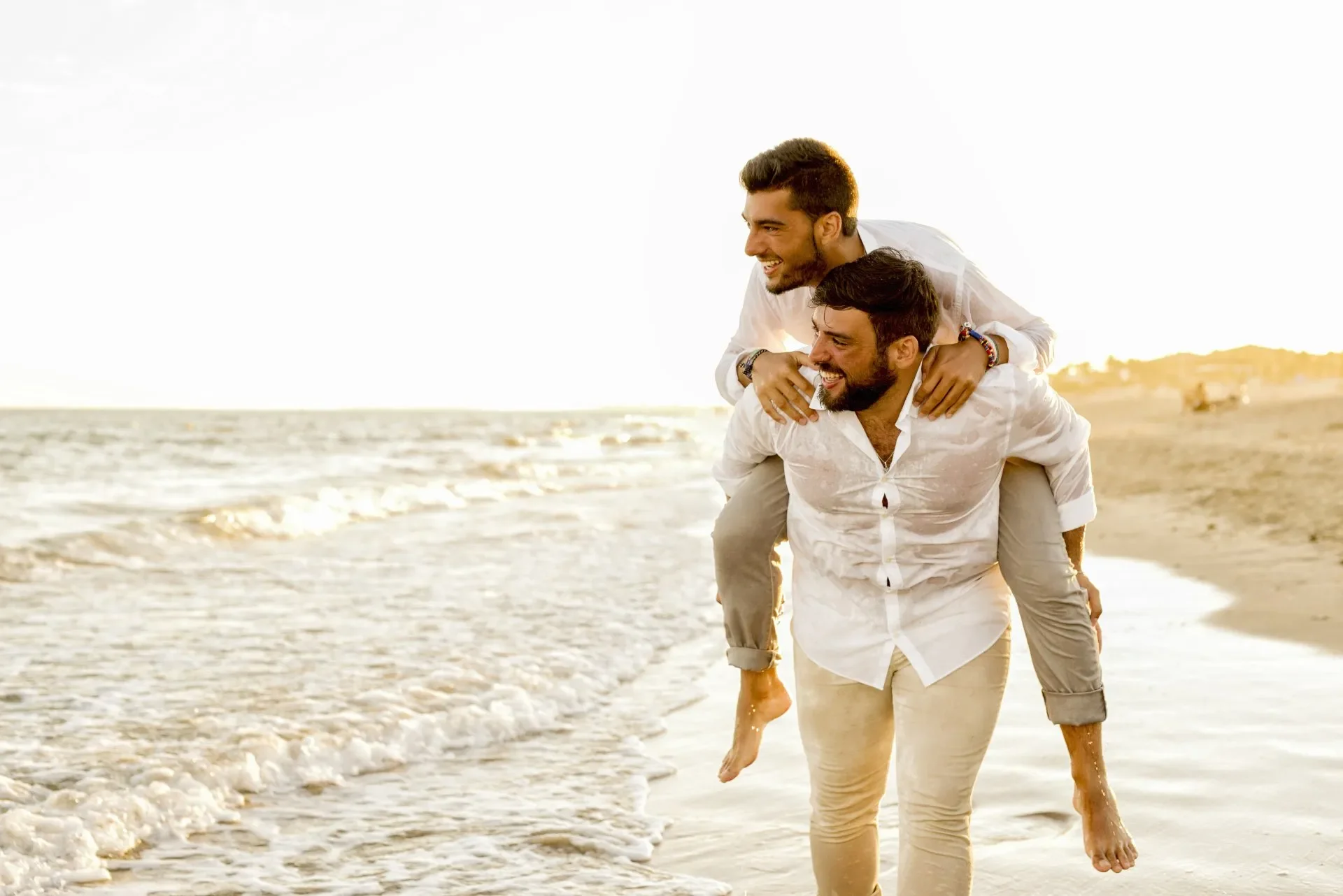 Two men in white shirts and beige pants playing on the beach, with one giving the other a piggyback ride during sunset.