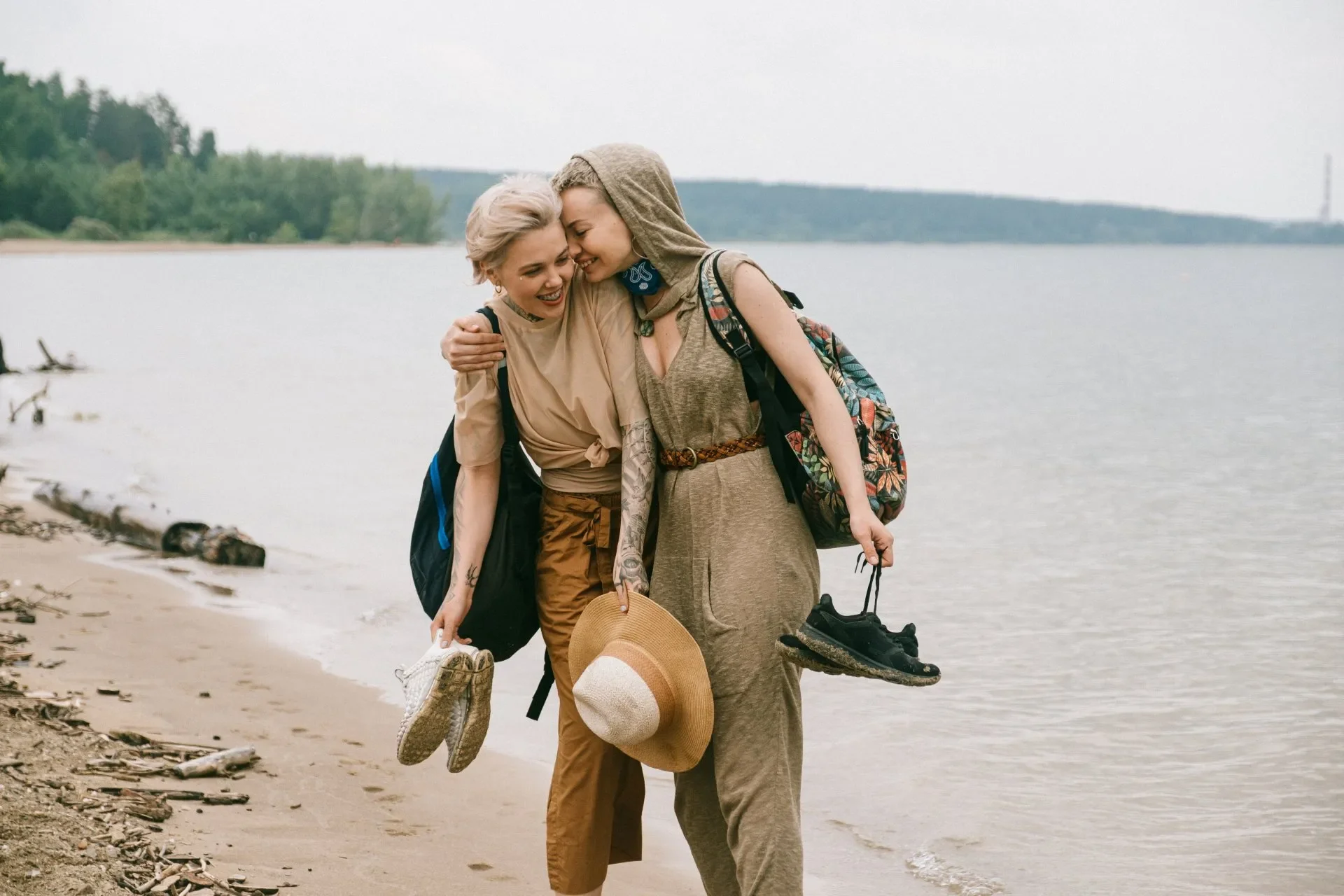 Two women happy and smiling, leaning into each other by a lakeshore, holding shoes and a hat, in a casual outdoor setting with trees and water in the background.