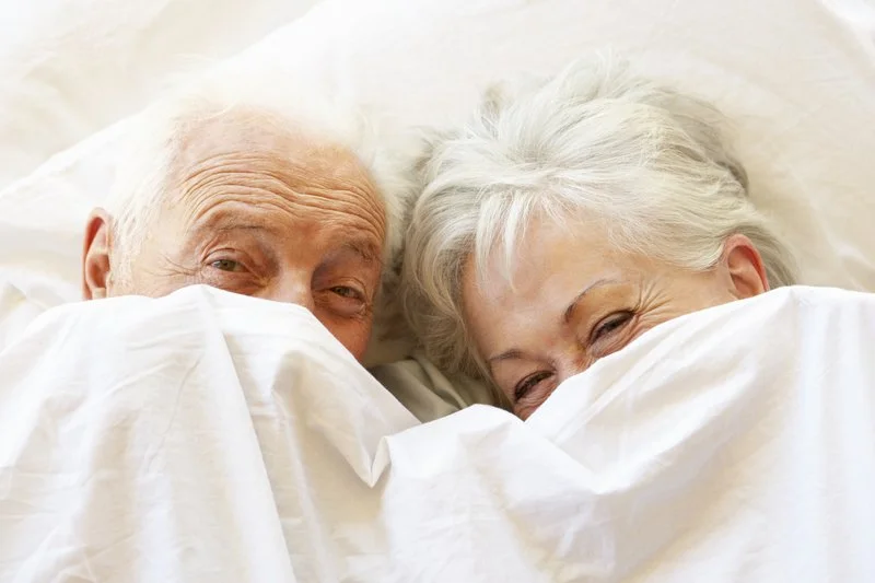 An elderly couple smiling and cuddling under a white blanket in bed.