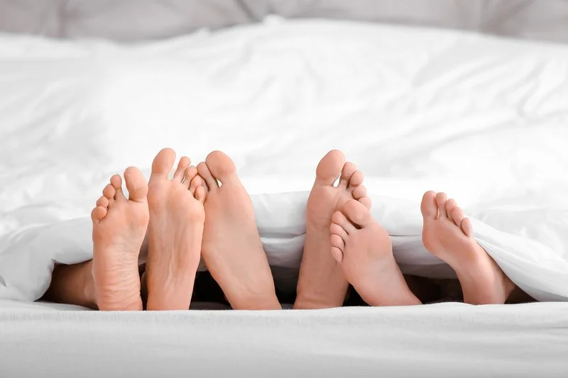 Multiple pairs of feet sticking out from under a white bedsheet on a bed.