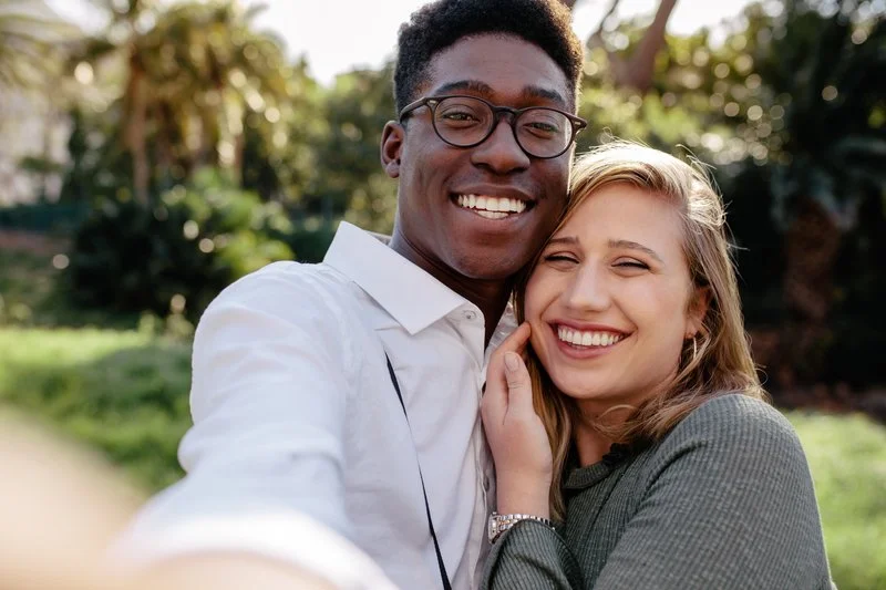 A young couple smiling and hugging outdoors, with greenery and trees in the background.