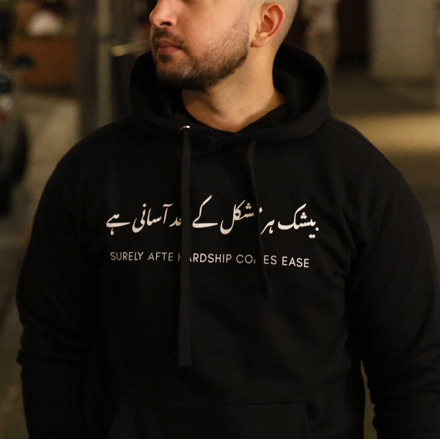 A man wearing a black hoodie with English and Urdu text on it, standing outdoors at night.