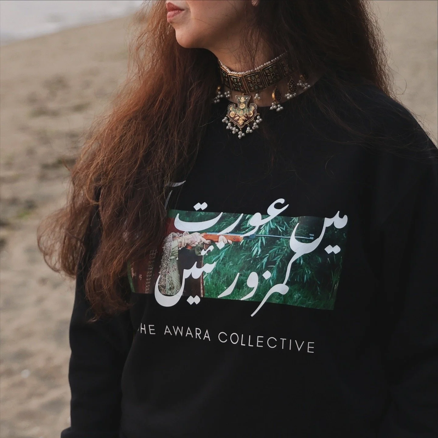 A person with long, wavy hair wearing a black shirt featuring a colorful graphic and text in a native script, along with a patterned choker necklace and jewelry, standing outdoors on a sandy area.