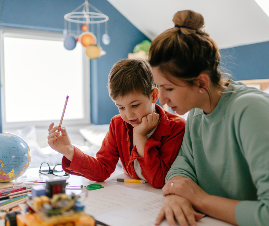 A woman and a boy studying together at a desk with school supplies, globe, and papers in a room with a blue wall and a window.