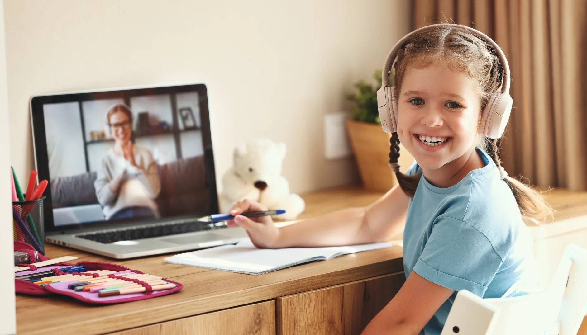 A young girl with braided hair smiling at the camera wearing headphones, sitting at a desk with a laptop and school supplies, while on a video call with a woman on the laptop screen.