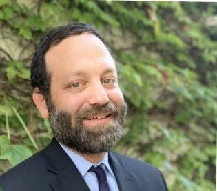 A man with a beard and dark hair, wearing a suit and tie, smiling outdoors with greenery in the background.