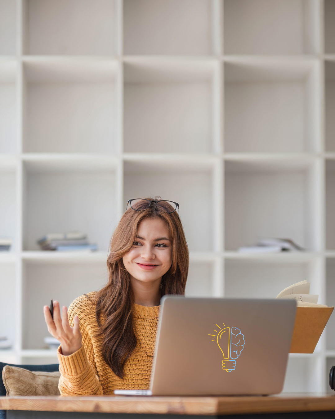 A young woman with long brown hair and glasses on her head, sitting at a desk with a laptop that has a light bulb with a brain illustration on it, holding a pen and a notebook, smiling in front of a white shelving unit.
