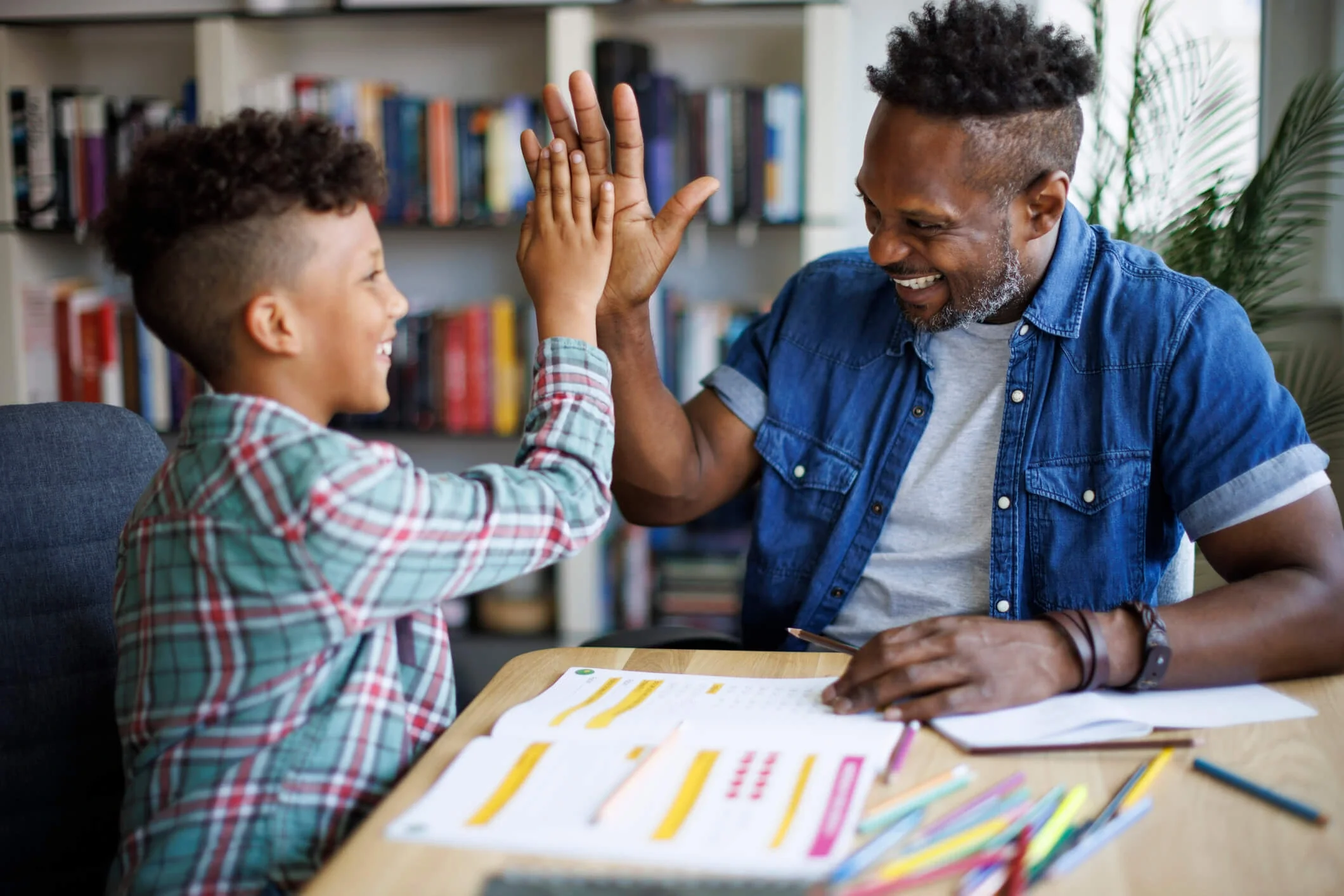 A young boy and an older man, possibly his father or teacher, giving each other a high five while sitting at a table with papers and colored pencils in a room with a bookshelf and a plant in the background.