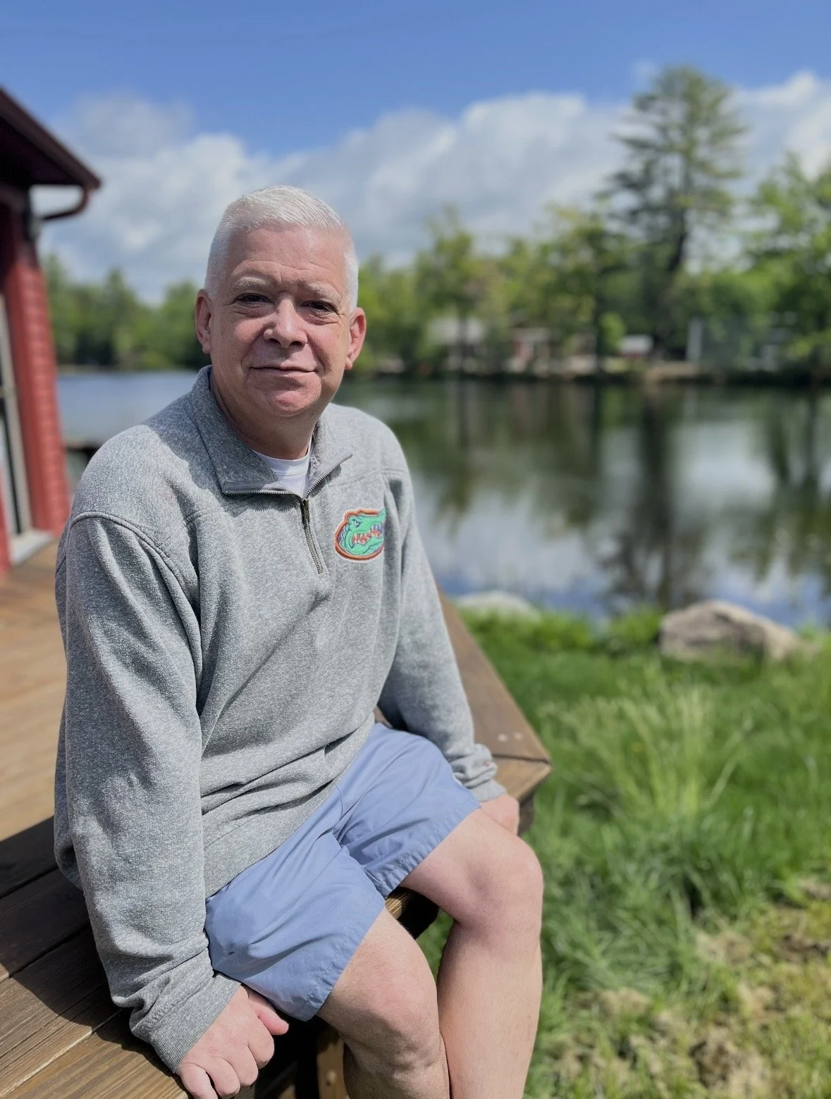 A man sitting on a wooden dock near a lake with trees in the background, wearing a gray quarter-zip sweatshirt with a Florida Gators logo and light blue shorts on a sunny day.