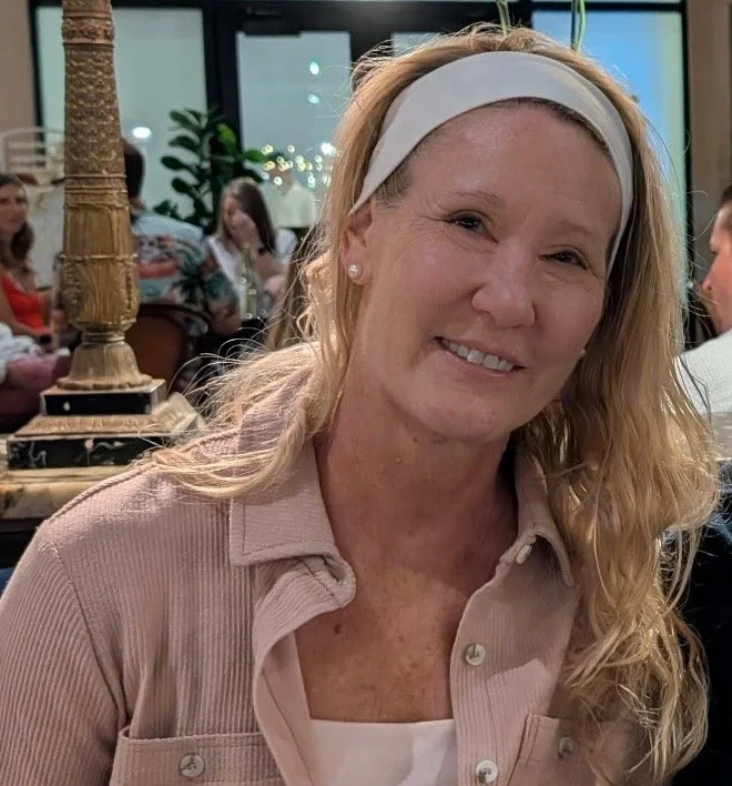 A middle-aged woman with long blonde hair, wearing a white headband and a light pink shirt, smiling at the camera in a restaurant or cafe setting.