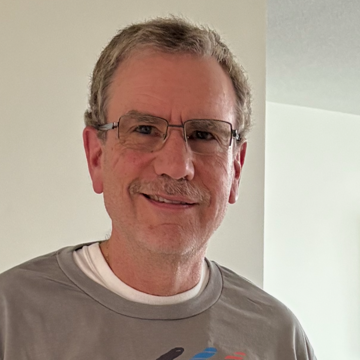A middle-aged man with glasses and short hair, smiling, wearing a Snoopy tennis team T-shirt with the text "1950 Peanuts Tennis Team."