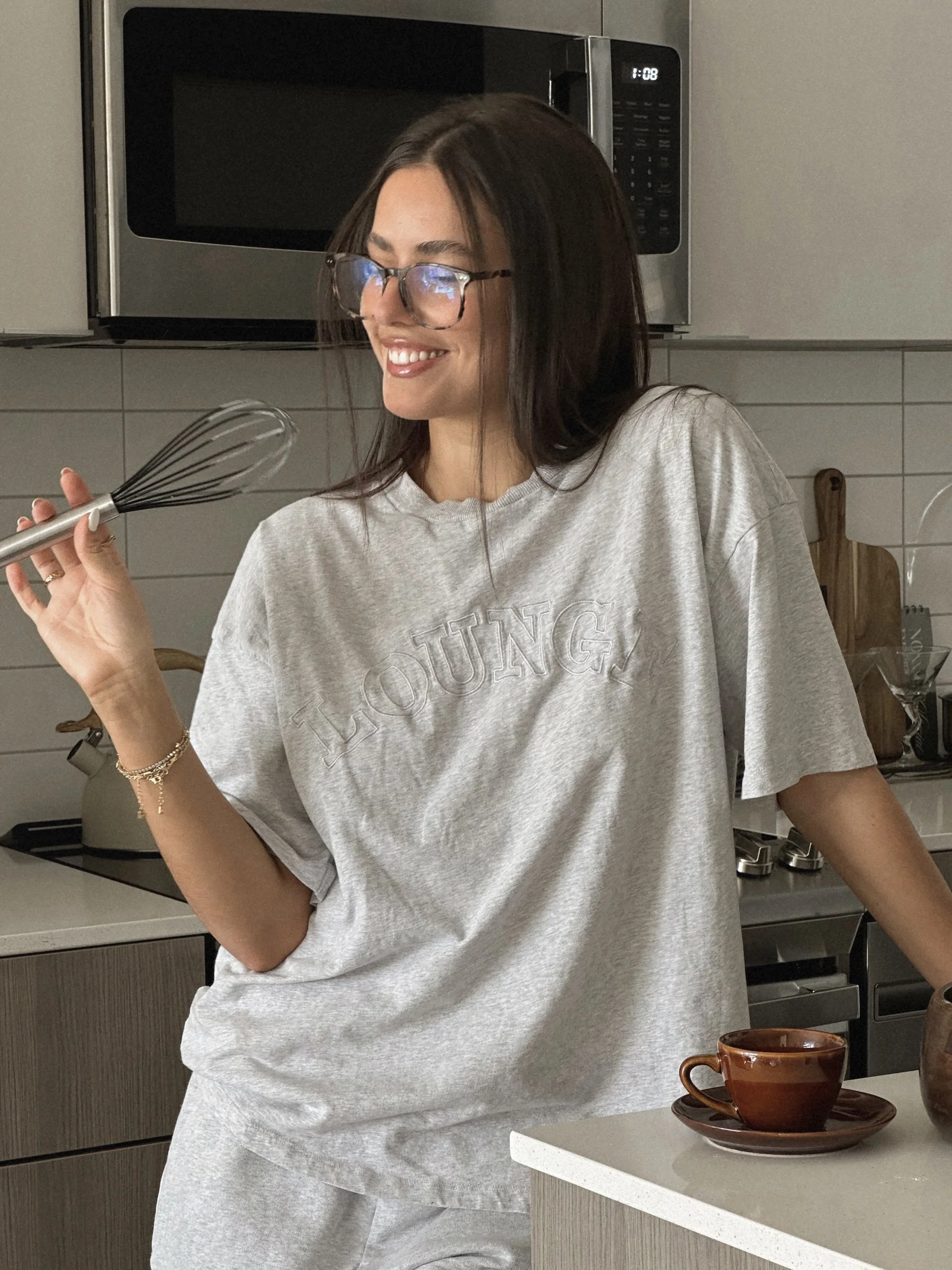 A young woman with long dark hair, wearing glasses and a gray T-shirt, is smiling and holding a whisk in a kitchen with a microwave, cups, and a cutting board visible in the background.