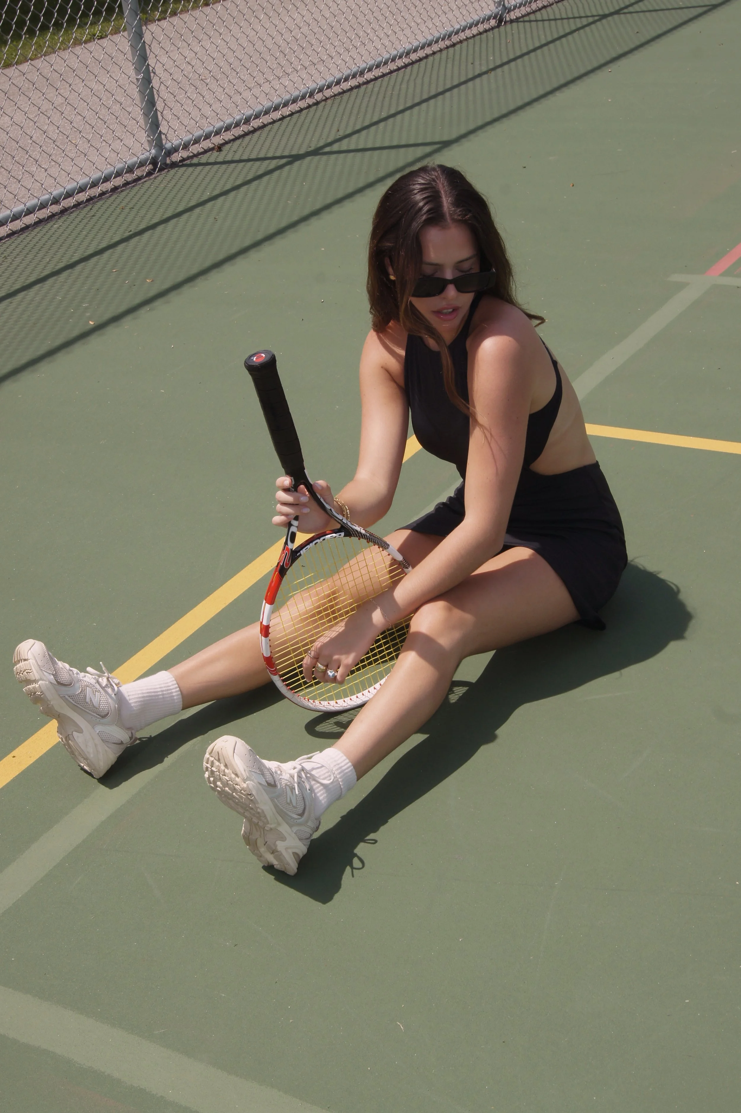 A woman sitting on a tennis court with her legs extended, holding a tennis racket, wearing sunglasses, a black tank top, black shorts, white sneakers, and white socks.