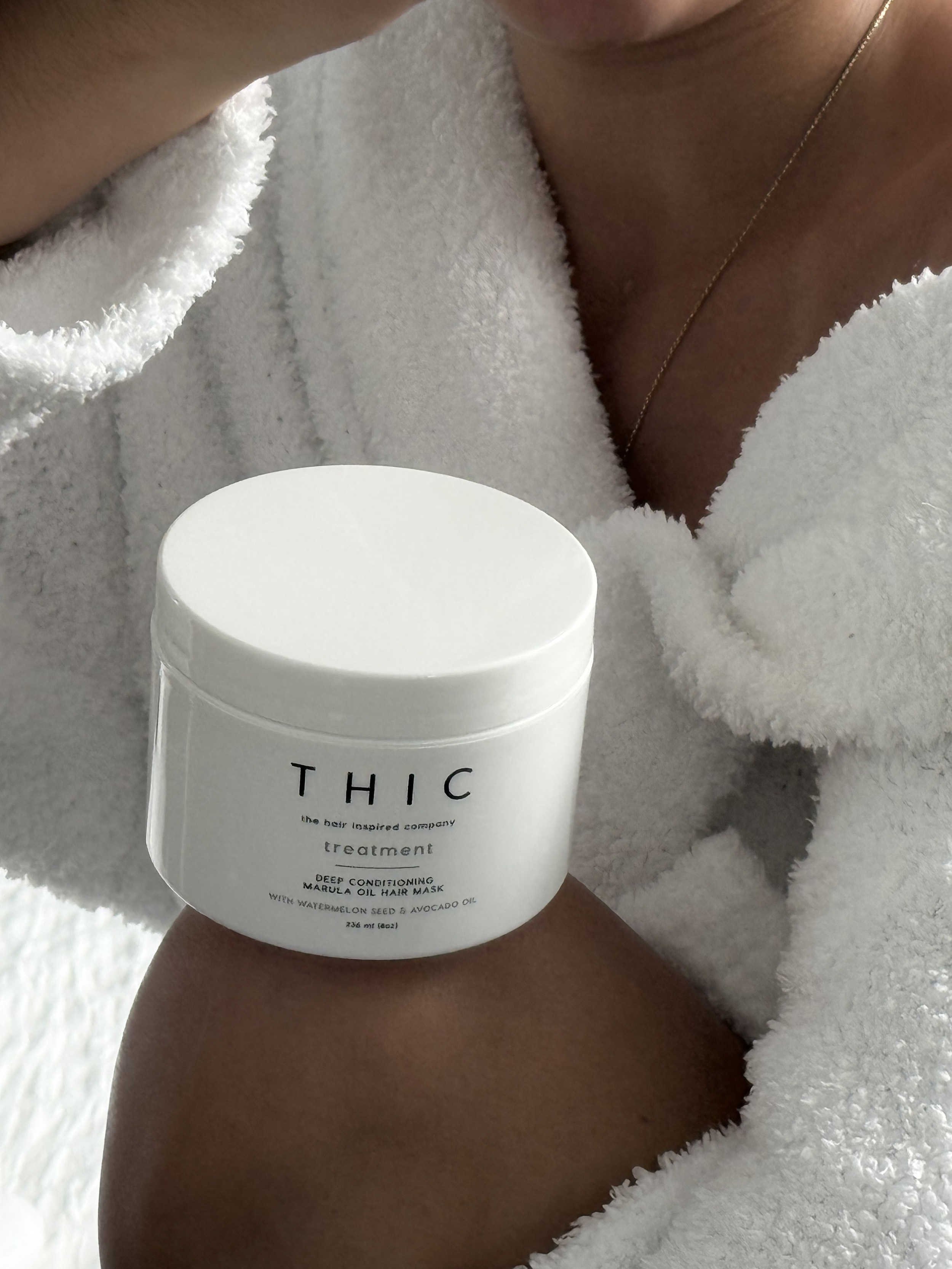 Person wearing a white fluffy robe holding a white container labeled 'THIC treatment deep conditioning marula oil hair mask' with watermelon seed and avocado oil.