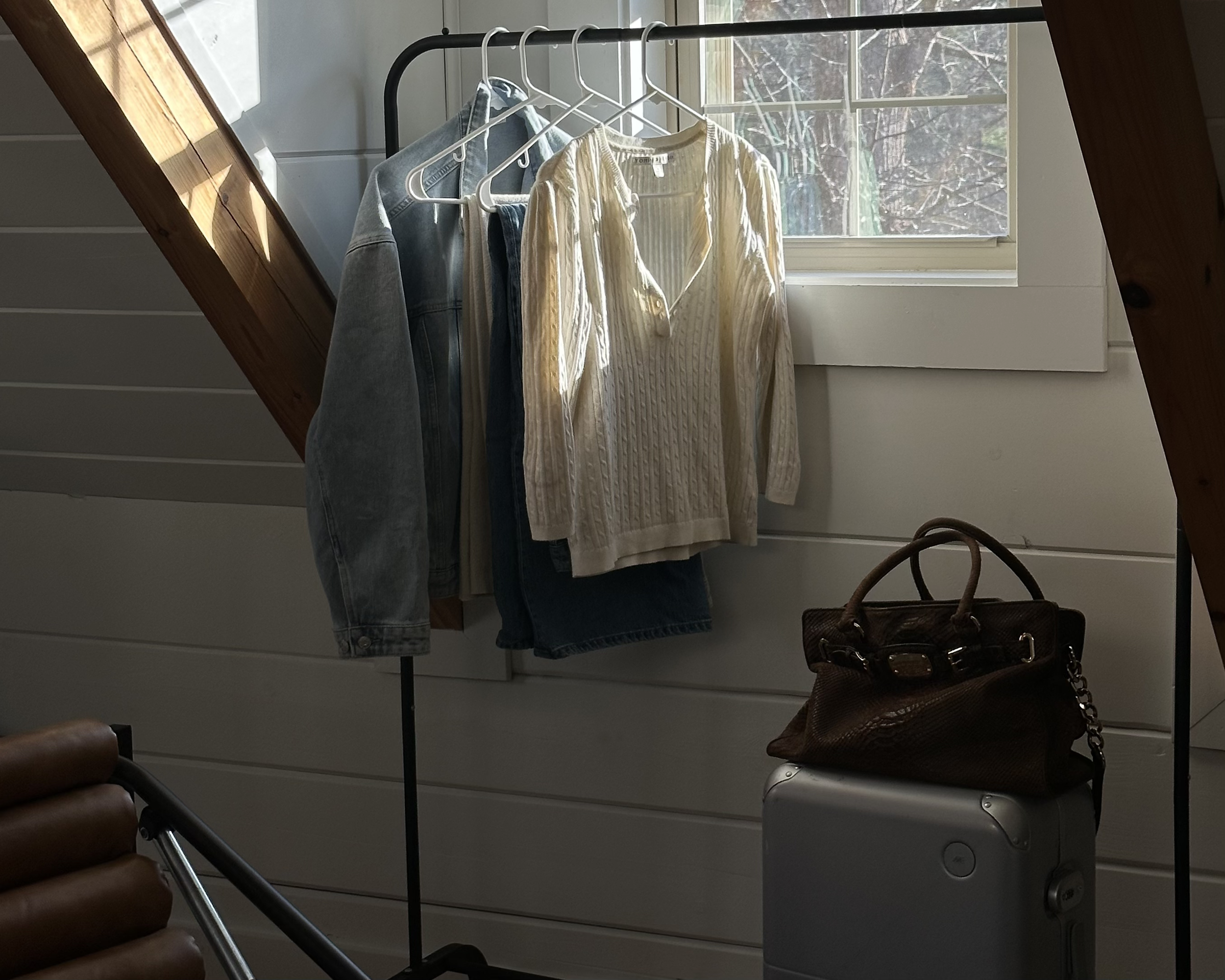 Clothes hanging on a portable clothing rack near a window, with a brown handbag and silver suitcase placed below the rack.