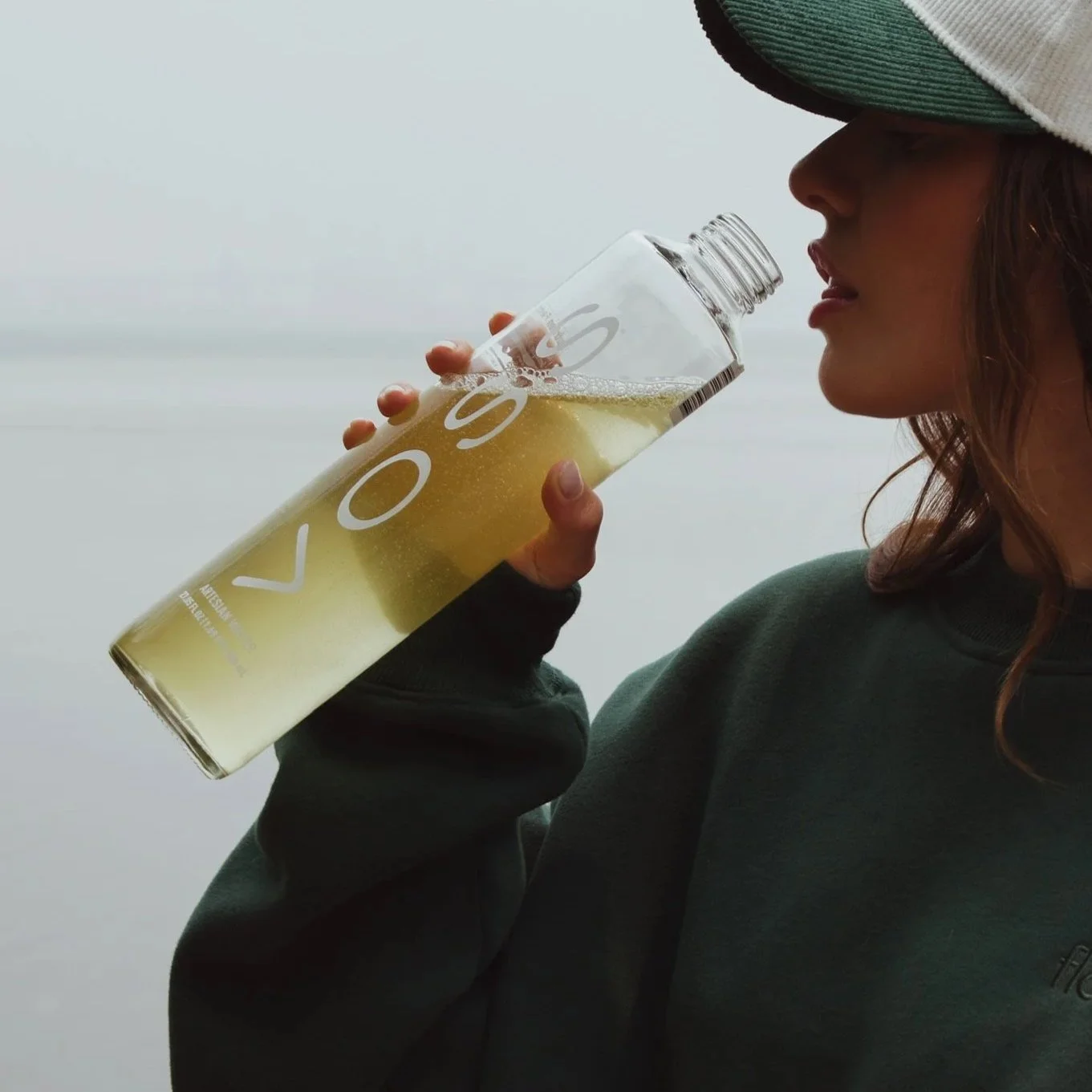 A woman wearing a hoodie and a cap is holding and drinking from a tall glass of pale yellow beverage with bubbles, with the letters 'VOSS' printed on the glass.