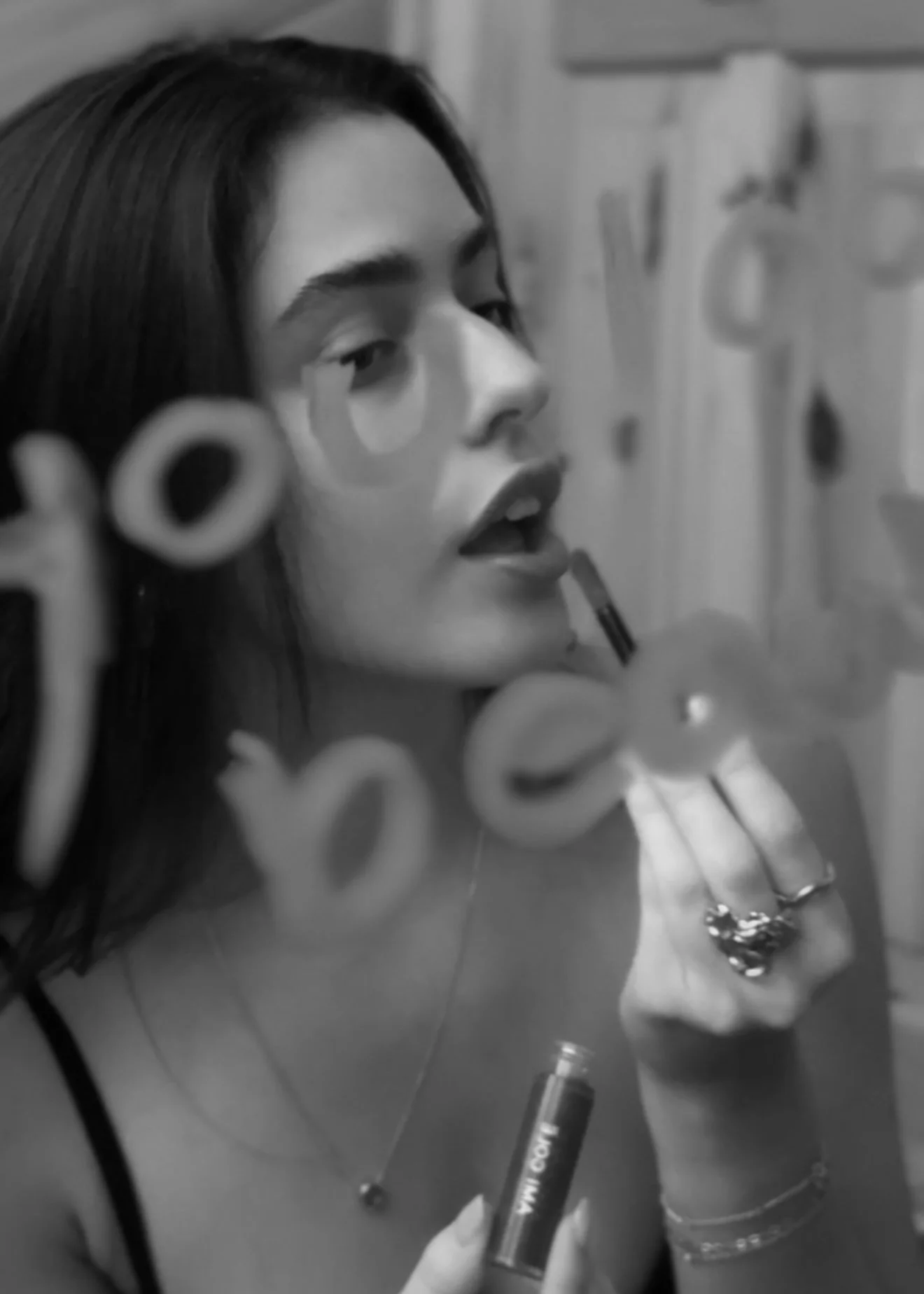 A young woman with dark hair, applying lipstick while looking into a mirror with writing on it. She's wearing rings, bracelets, and a necklace.
