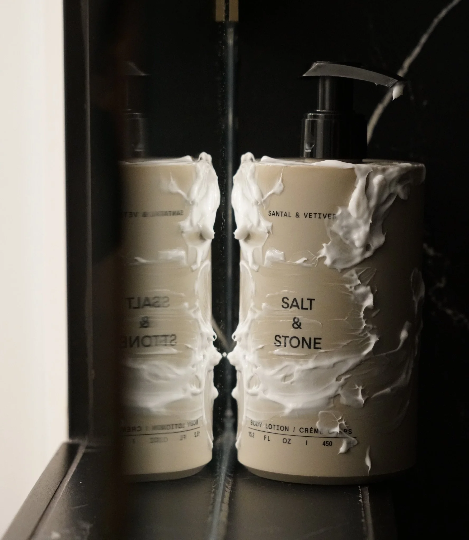 A mirror reflecting a beige bottle of Salt & Stone body lotion with a black pump, with lotion spilled on the bottle, set against a dark background.
