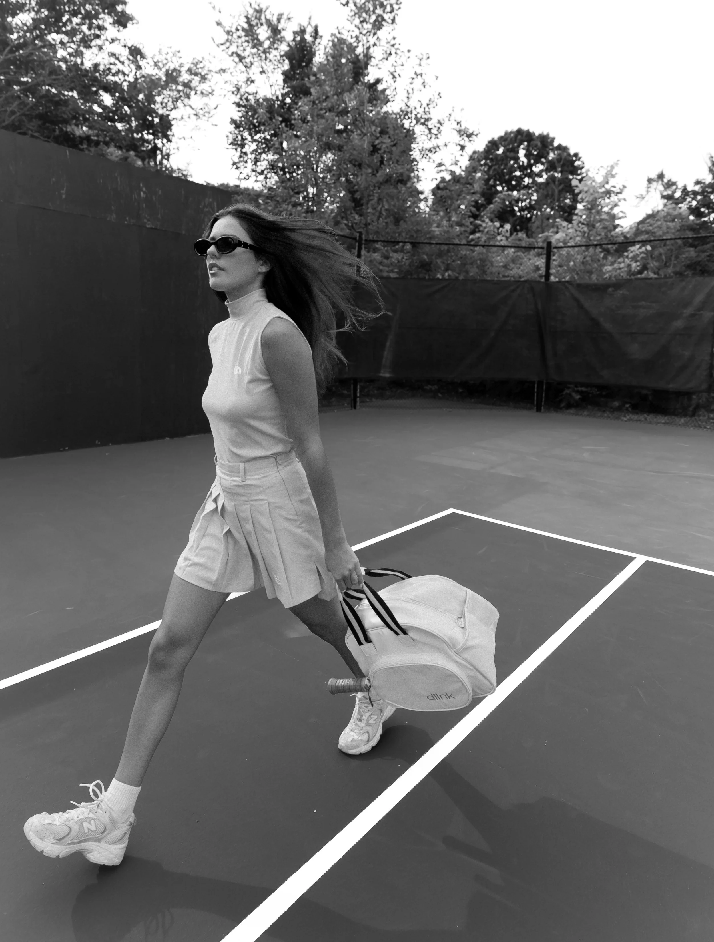 A woman walking on a tennis court, carrying a tennis bag, wearing sunglasses, a sleeveless top, shorts, and athletic shoes, with trees in the background.